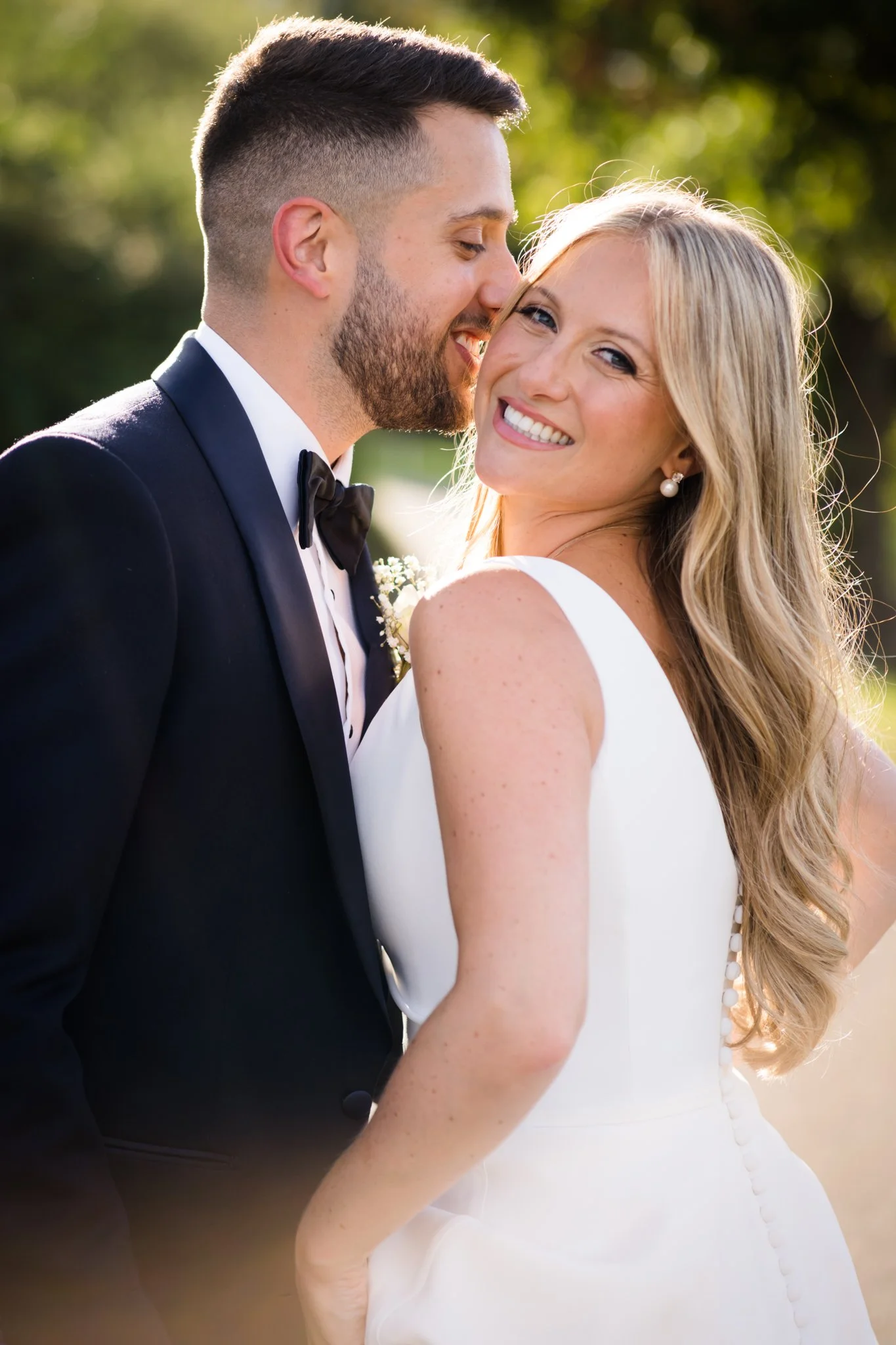 A newlywed couple smiling and posing outdoors, the groom wearing a tuxedo and the bride in a white wedding dress, with sunlight in the background.