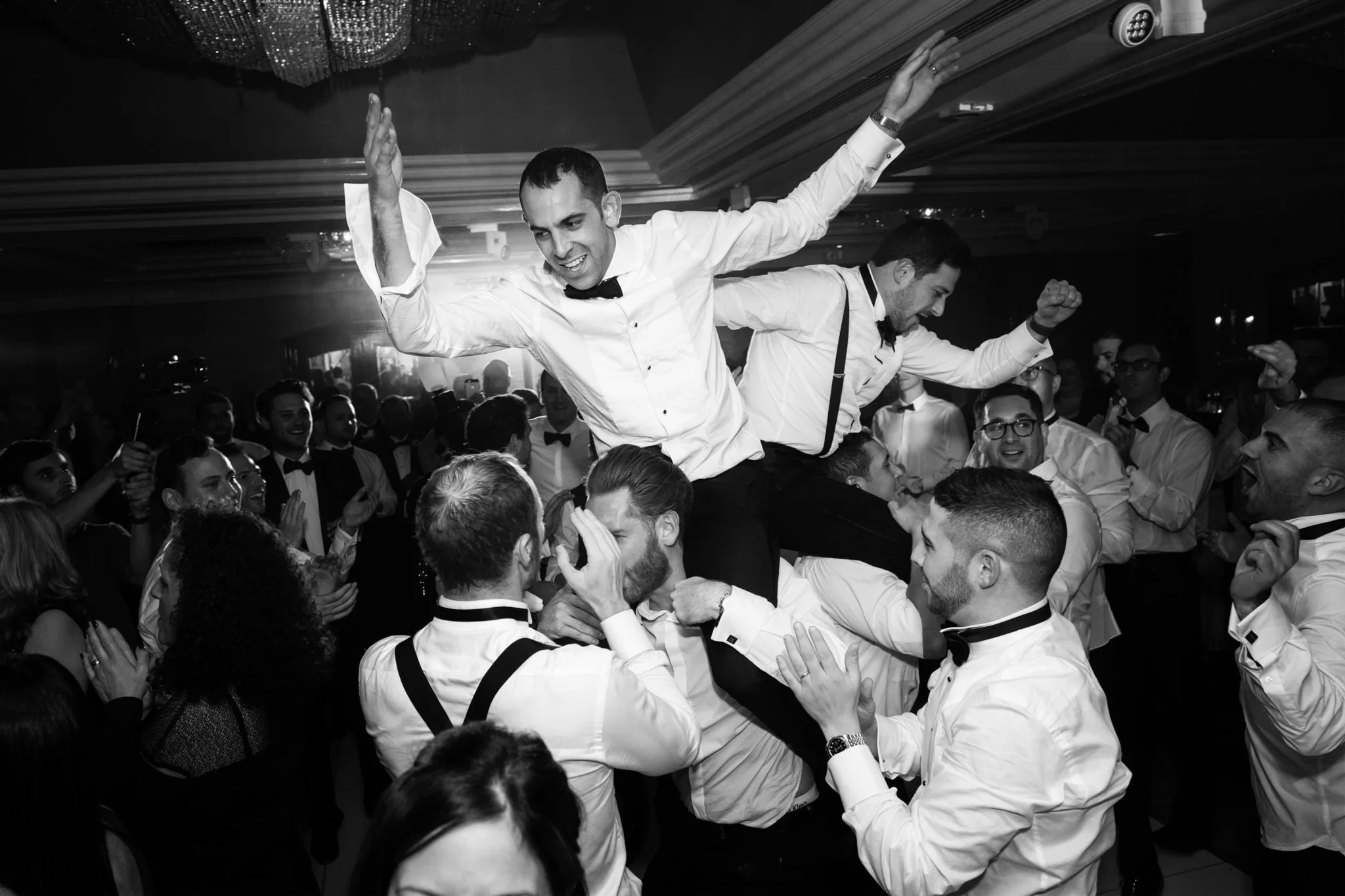 People celebrating at a wedding reception, with two men lifted on others' shoulders, dancing and smiling.