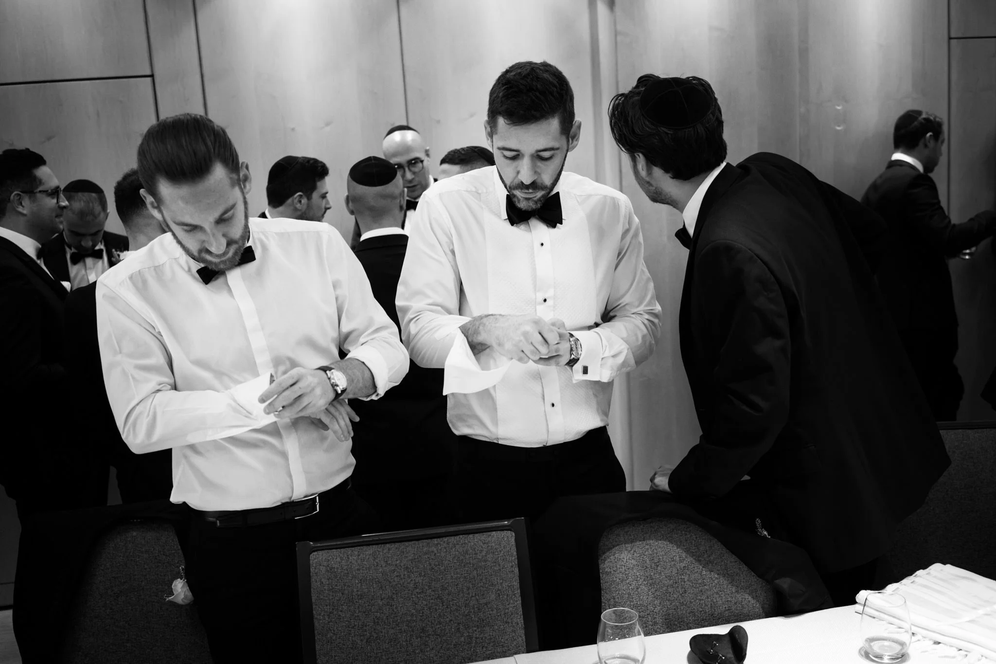 Three men dressed in formal attire with bow ties, preparing for a formal event, with others in similar attire in the background, in a room with wood-paneled walls.
