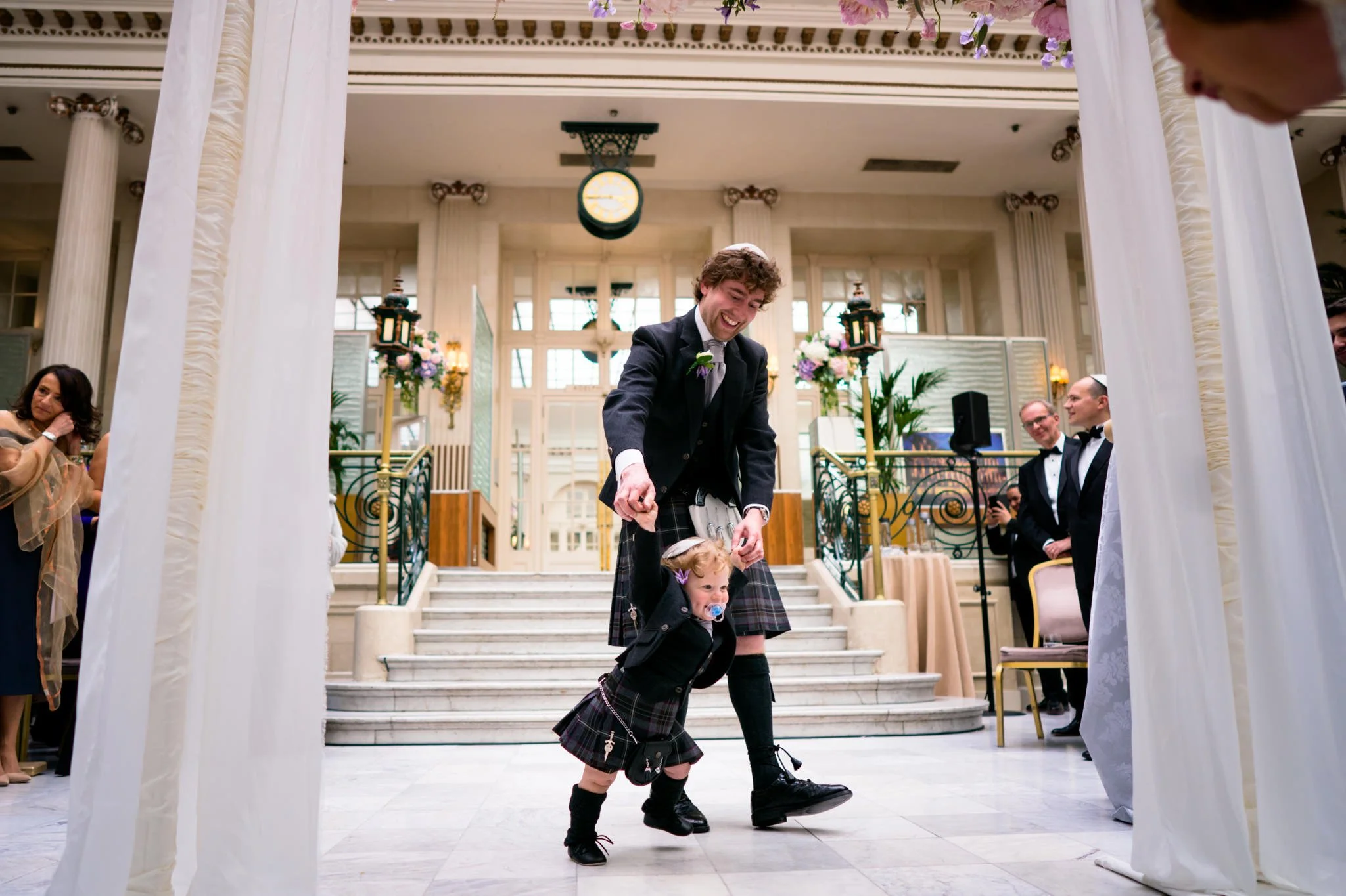 A young man in traditional Scottish attire, including a kilt, is smiling and dancing with a young girl who is holding his hand. They are inside a grand, decorated venue with marble floors, staircase, and flowers, at a wedding or celebration event.