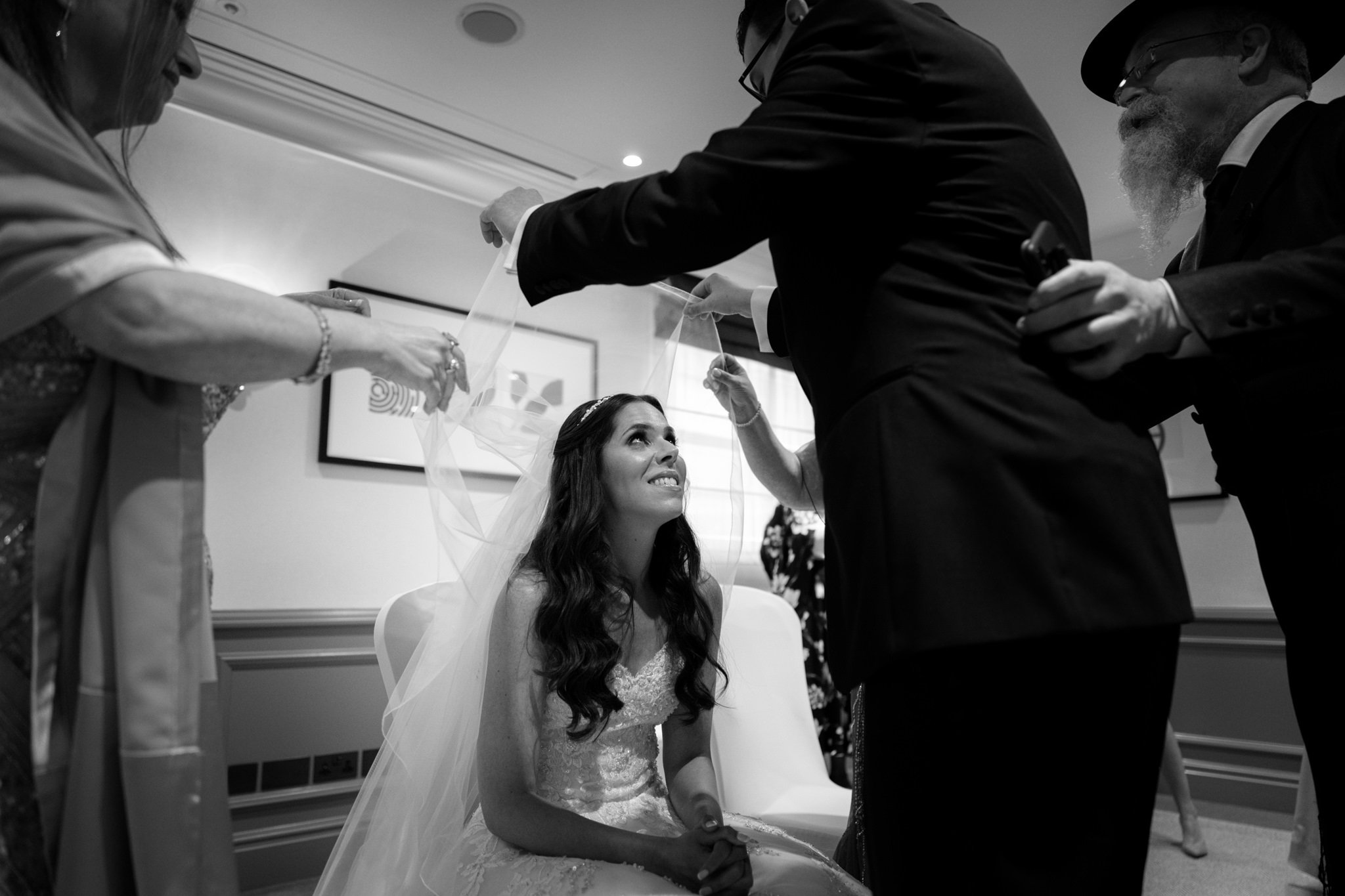 A bride sitting on a chair, smiling as she looks up, while family members help her with her wedding veil during a wedding ceremony.