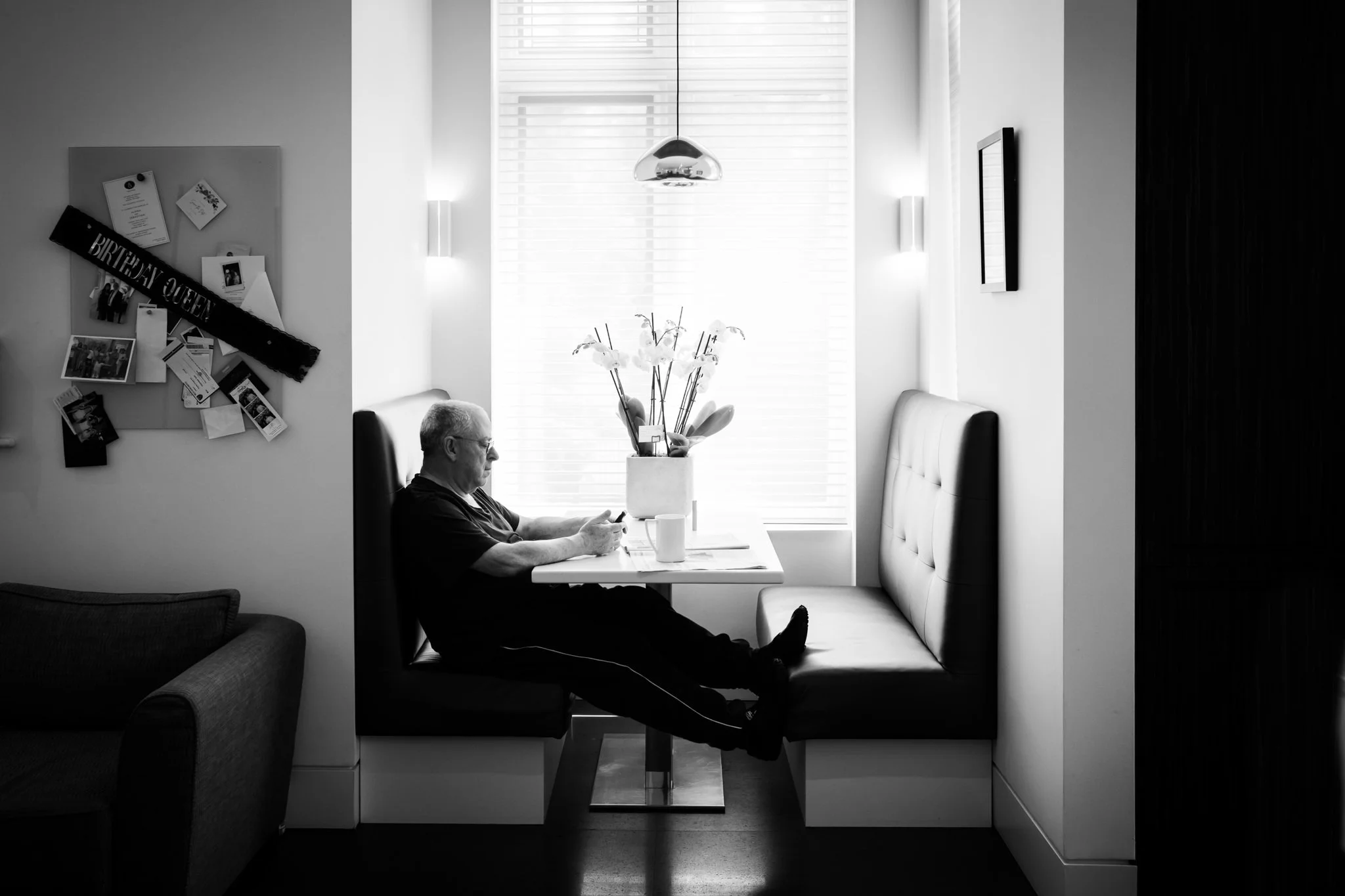 A black-and-white photo of an elderly man sitting in a booth at a cafe, looking at his phone. The booth has a high backrest, and there is a window with blinds behind him. A vase with orchids sits on the table in front of him, and a bulletin board wit