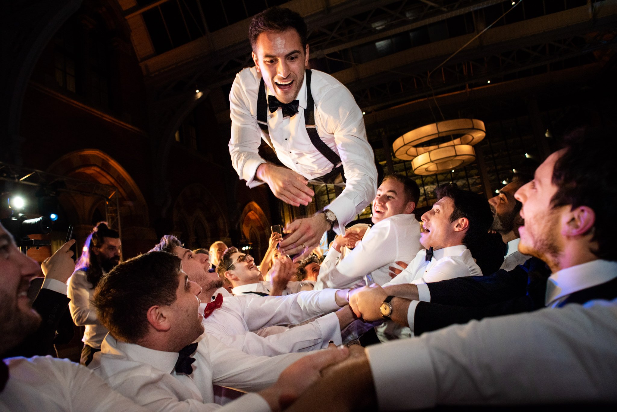 A man in a tuxedo is being lifted and celebrated by a group of men dressed in formal attire at a party or event.
