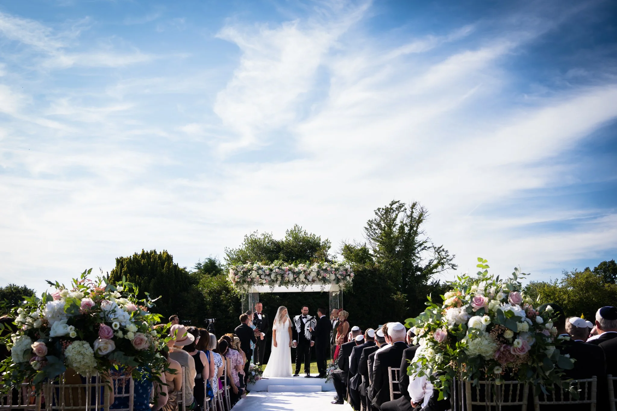 Outdoor wedding ceremony with a bride, groom, officiant, and guests seated on either side, under a floral arch, on a sunny day with blue sky and clouds.