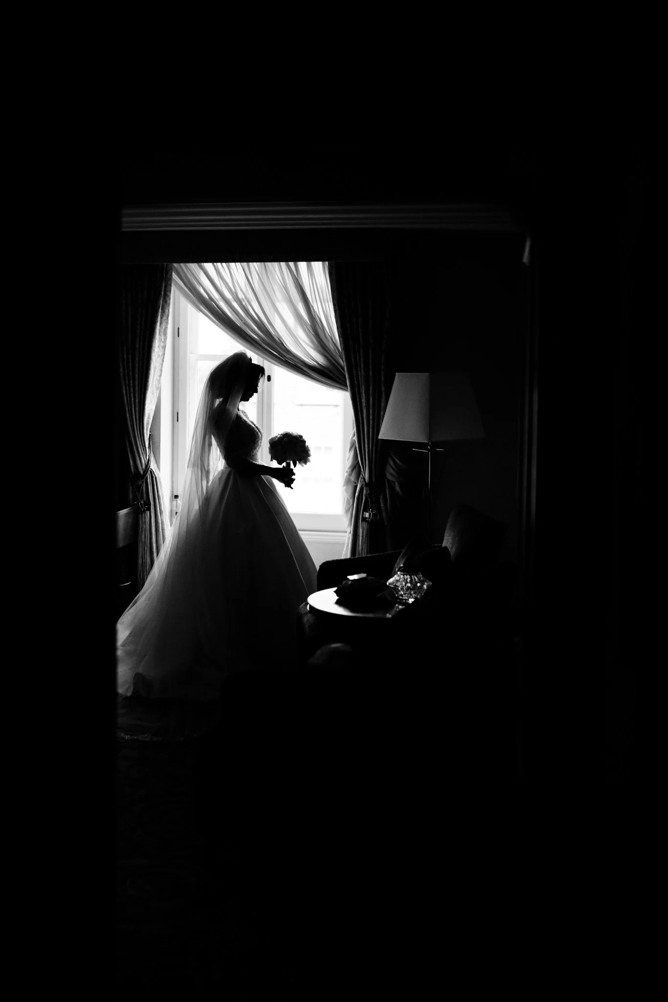 Silhouette of a bride in a wedding dress and veil holding a bouquet, standing by a window with curtains, in a dimly lit room.