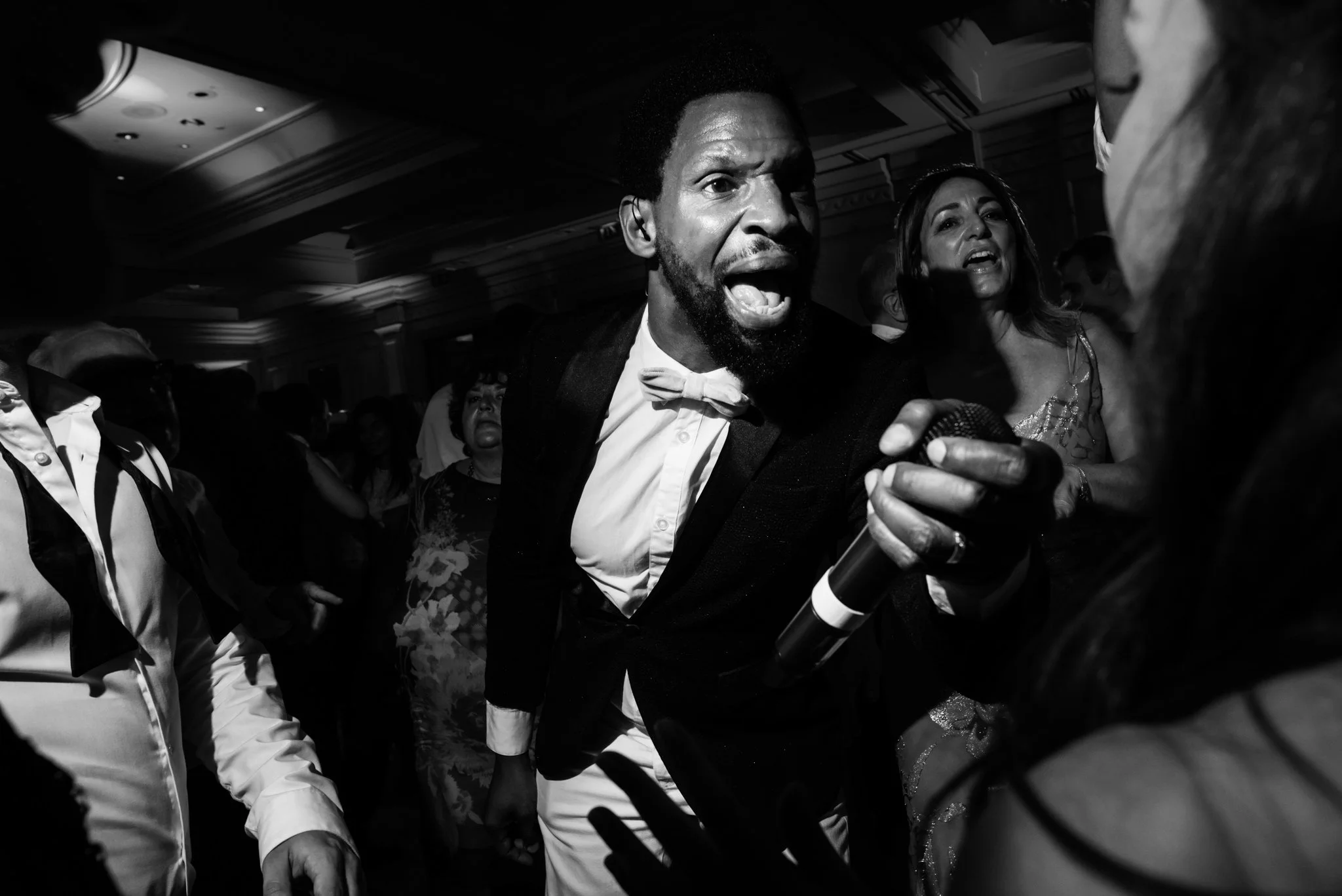A man dressed in a tuxedo with a bow tie holding a microphone appears to be speaking or singing passionately at a lively event or party, with several people around him in a spaced, indoor setting.