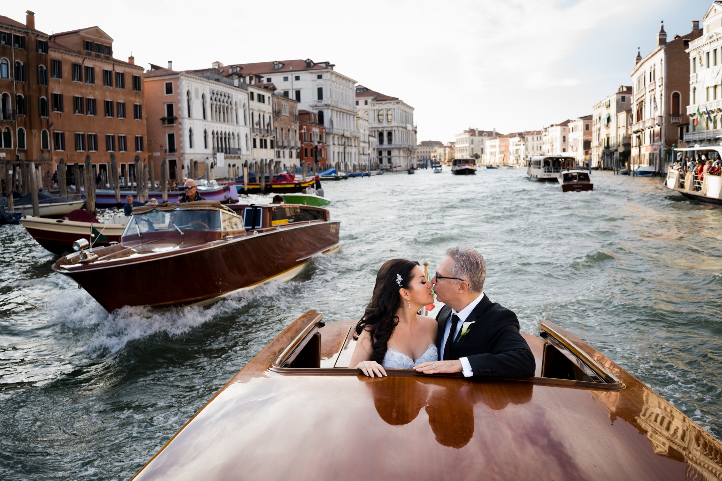A newlywed couple kiss on a wooden boat cruising through a canal in Venice, Italy, with historic buildings and other boats along the waterway.