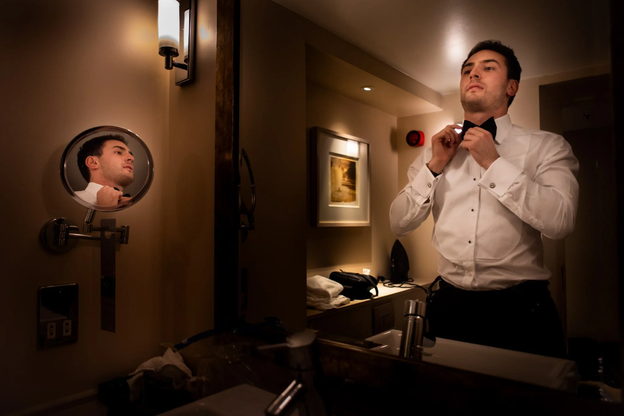 A man in a tuxedo getting ready in a hotel bathroom, adjusting his bow tie in front of a mirror.