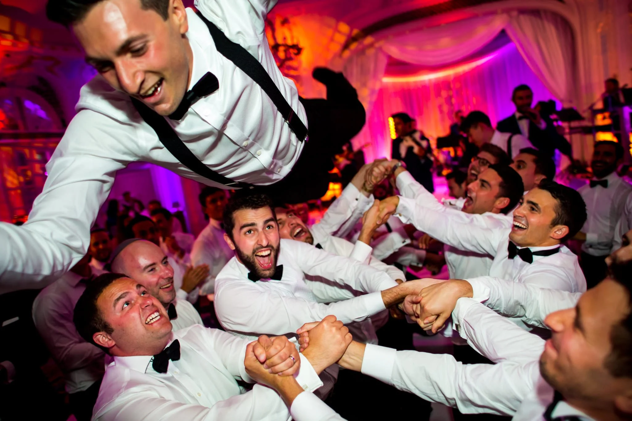Men in tuxedos dancing and celebrating at a wedding reception, with colorful lighting in the background.