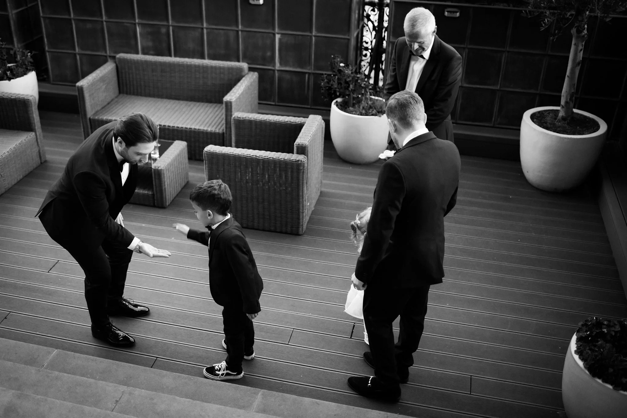 Adults and children dressed in formal attire on a wooden deck, with two large potted plants and wicker furniture, during a formal event or gathering.