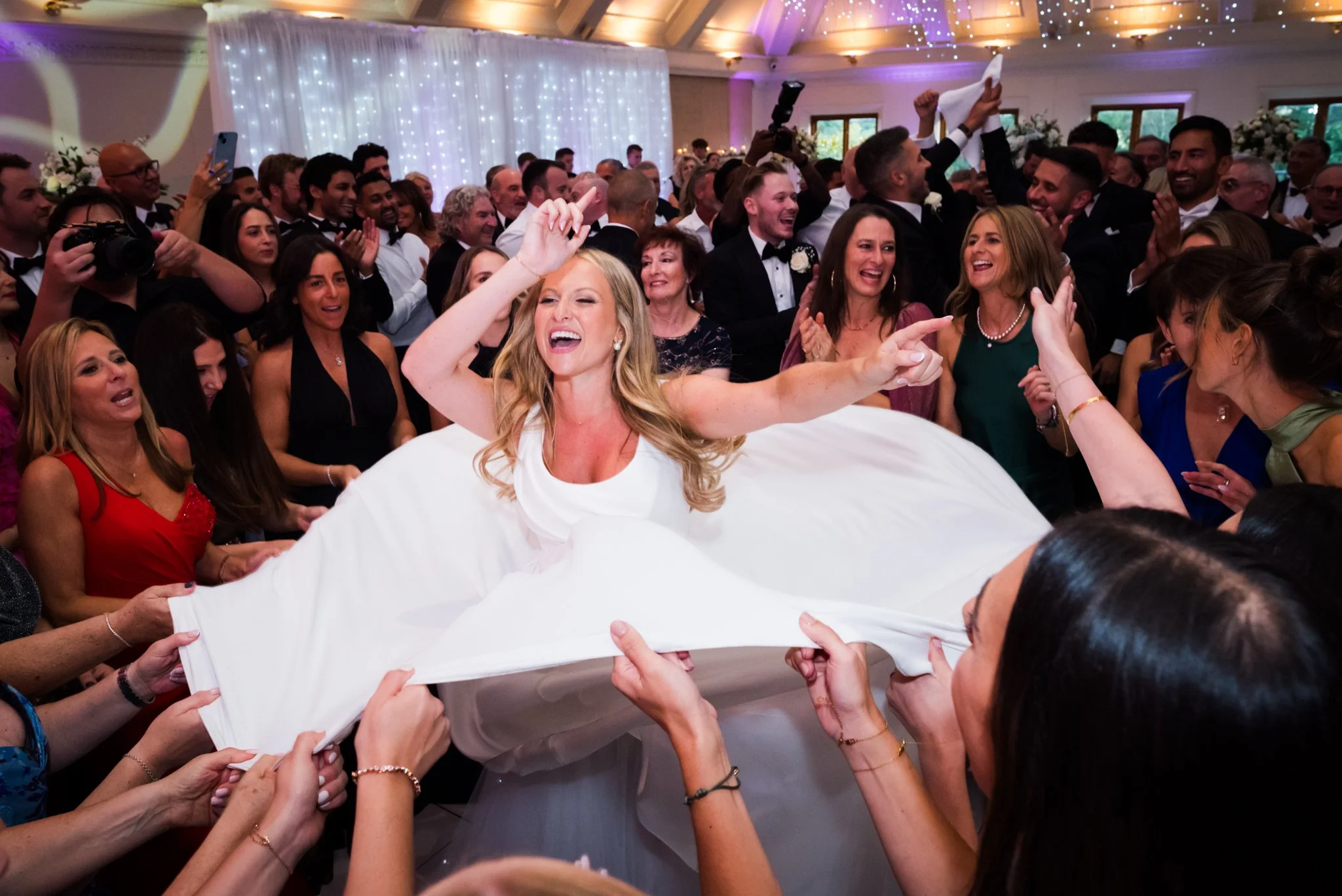 A woman in a wedding dress is being lifted and celebrated at her wedding reception, surrounded by smiling guests.