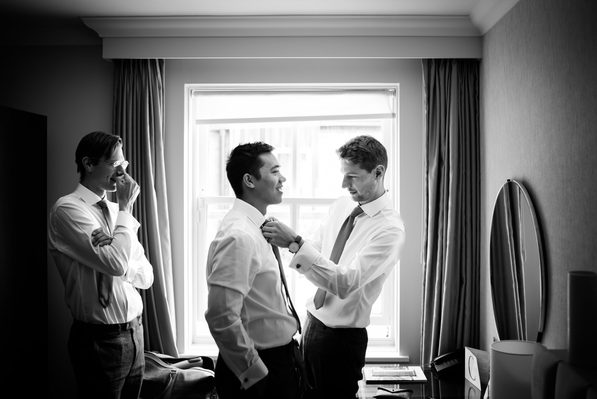 Three men in a hotel room, two of them are getting ready while one is smiling, and the third is adjusting the tie of the man in the middle, all dressed in white shirts and ties.