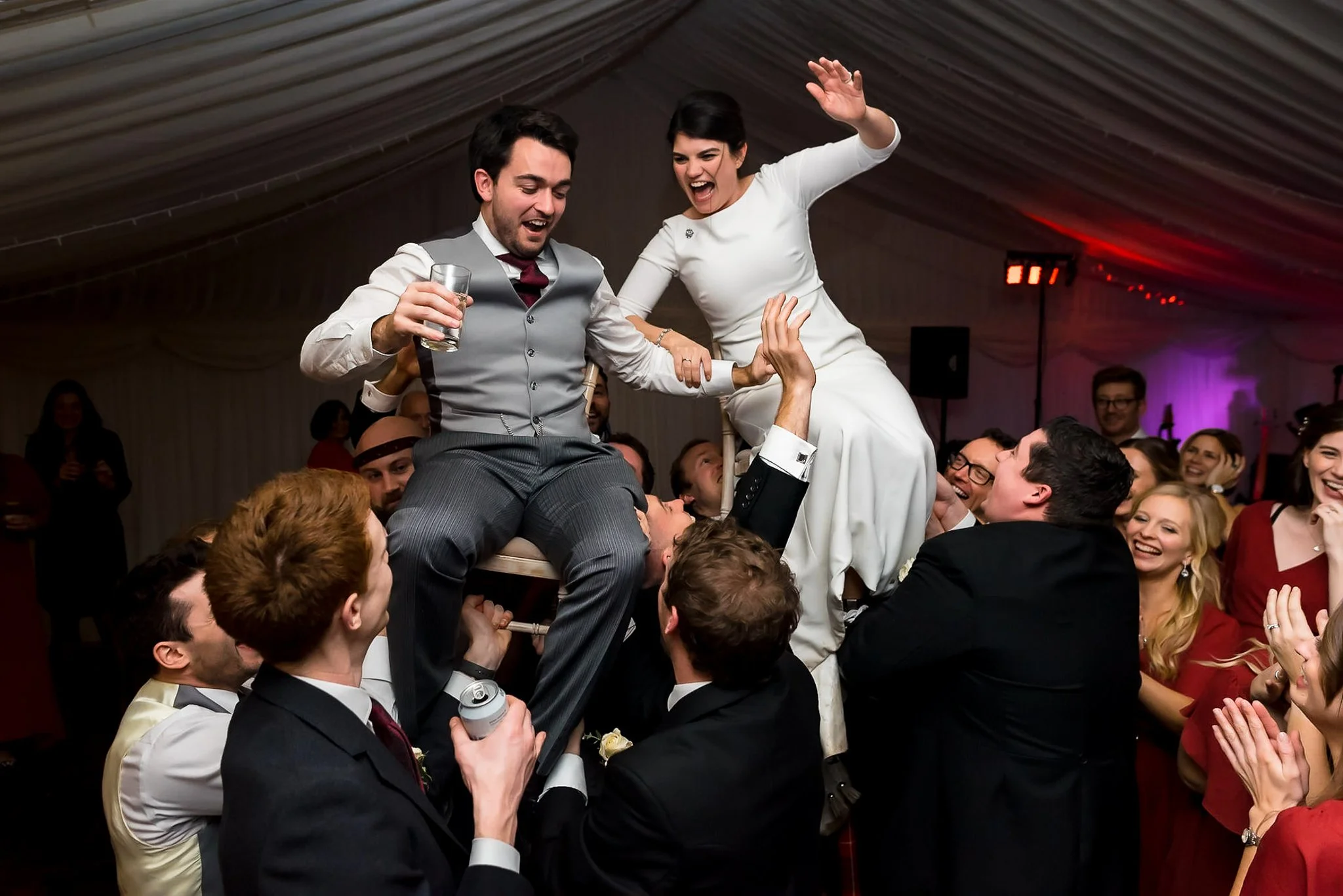 Wedding celebration with bride and groom sitting on a chair lifted by guests, surrounded by applauding friends.