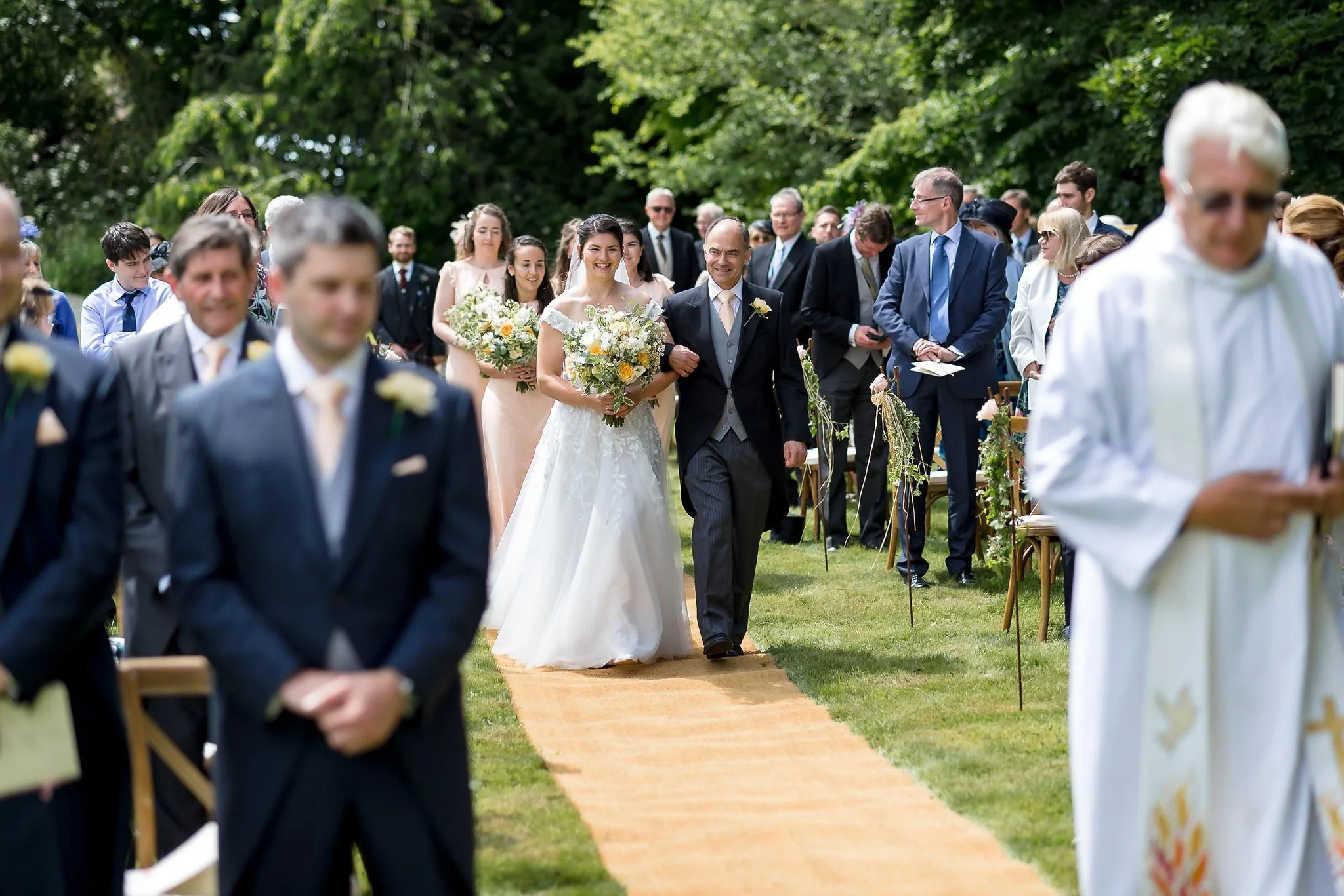 A bride walking down an outdoor wedding aisle on a sunny day, holding a bouquet, escorted by a man in a suit, with guests seated and standing on either side.