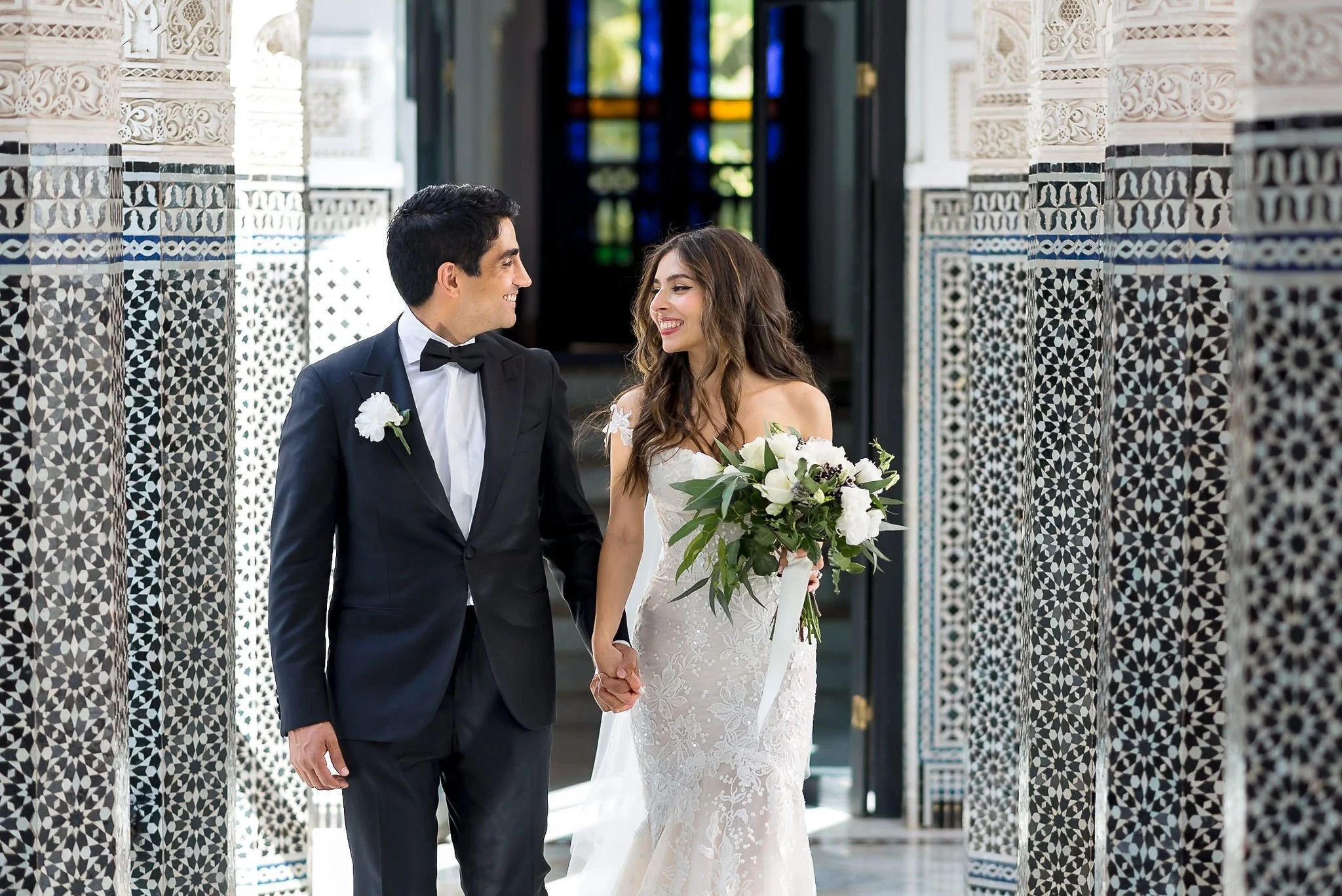 A bride and groom holding hands and smiling at each other inside a building with intricate patterned walls and a stained glass door in the background.