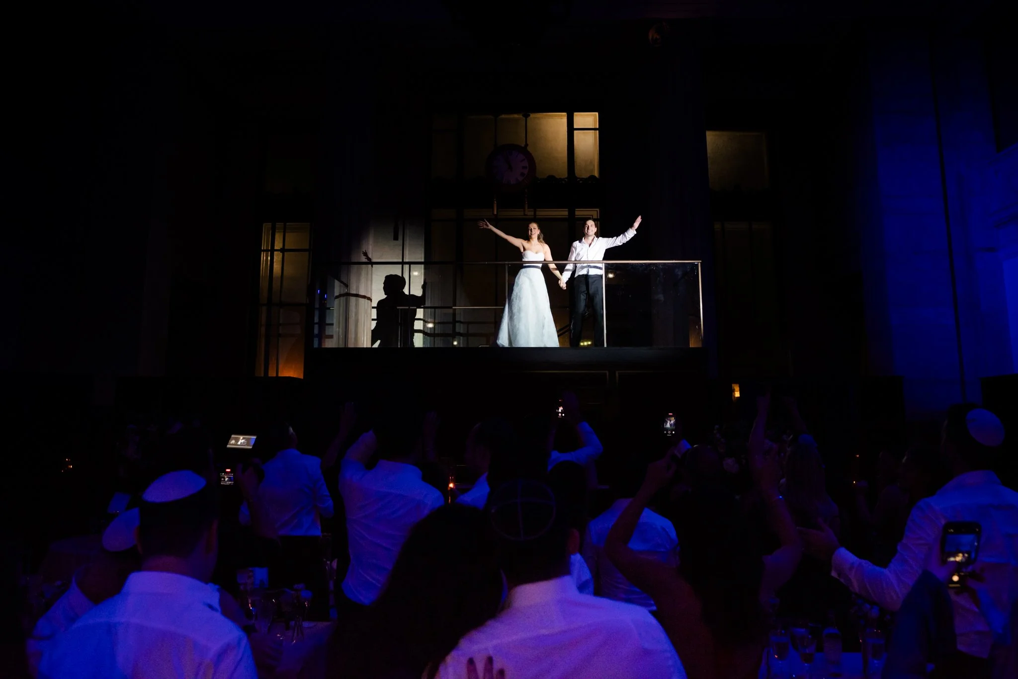 A bride and groom standing on a balcony holding hands during their wedding reception, with guests below taking photos and videos in a dimly lit hall.