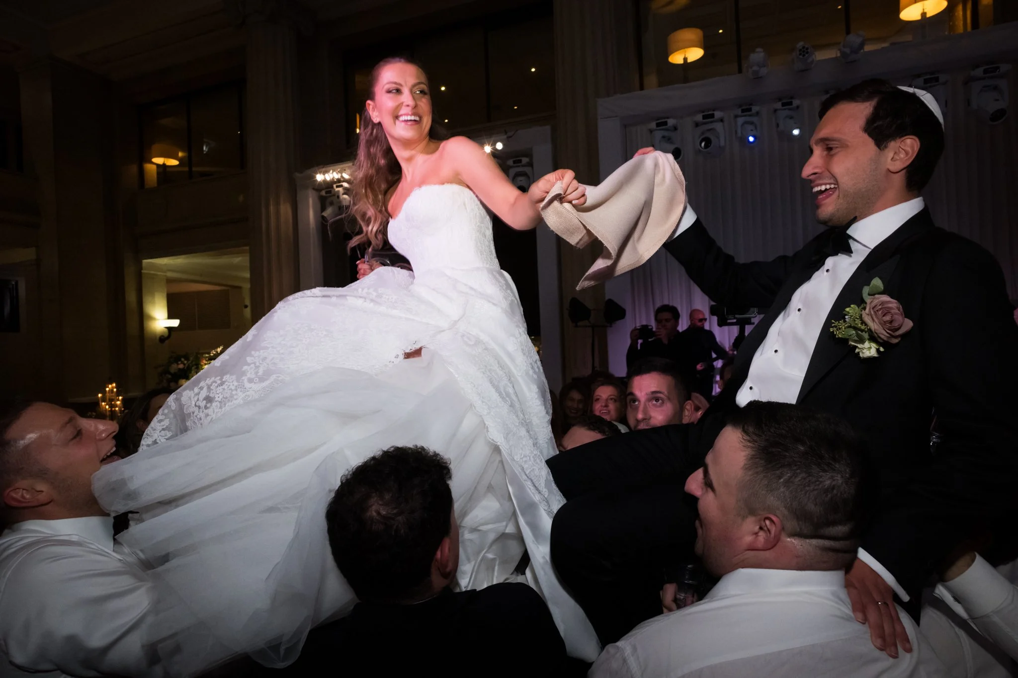 A bride and groom are being lifted in celebration at their wedding reception, with the bride smiling and holding a napkin.