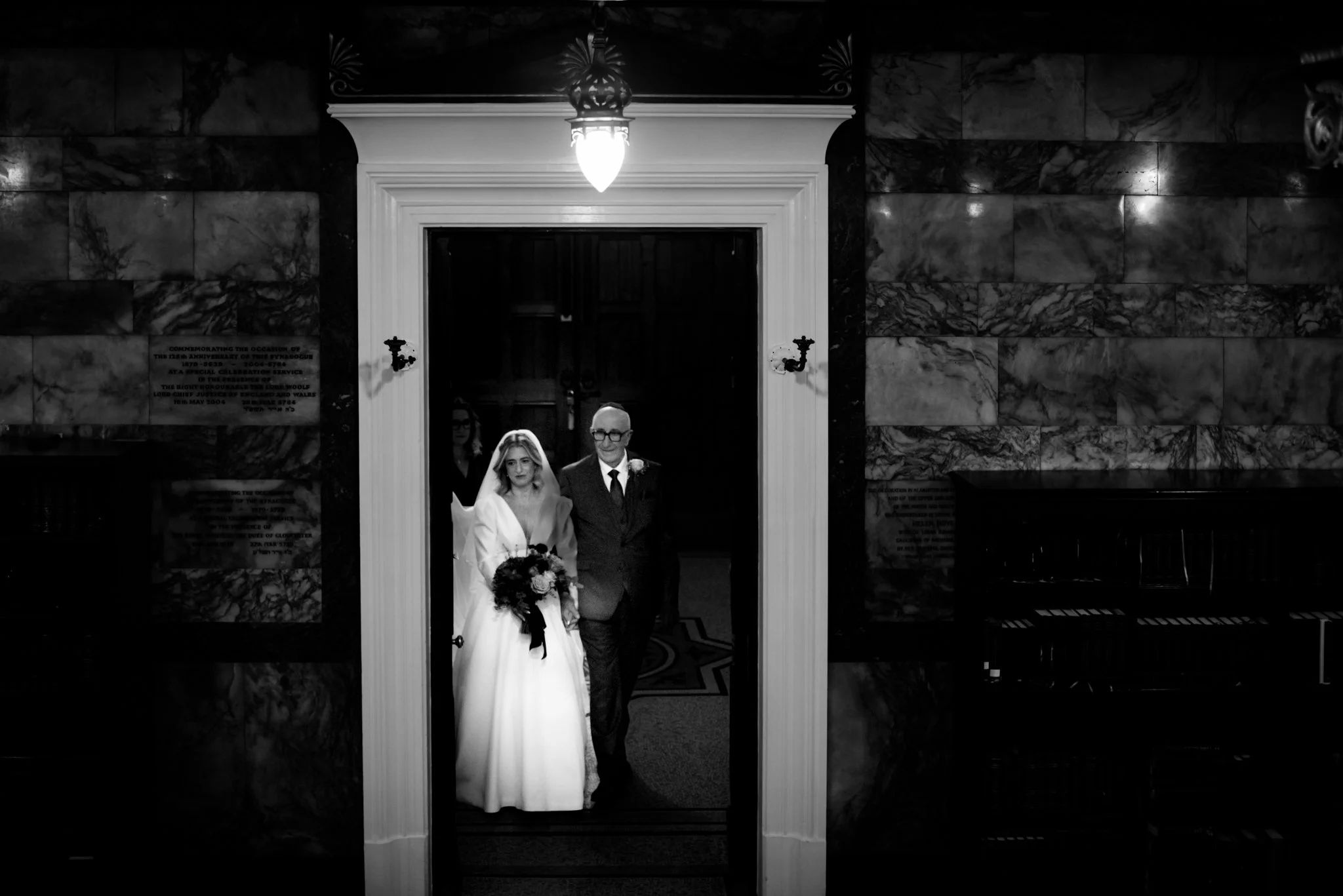 A bride in a wedding gown holding a bouquet walking with an older man in a suit through a doorway inside a building with marble walls, in black and white.