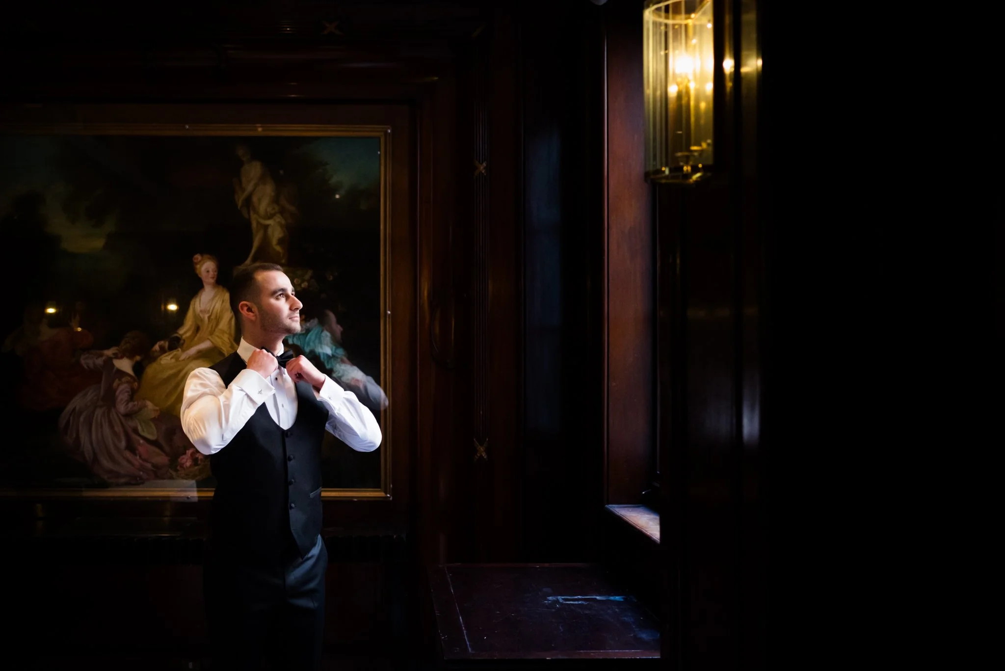A man in a tuxedo adjusting his bowtie in a dimly lit room with dark wooden walls and a large classical painting hanging behind him.