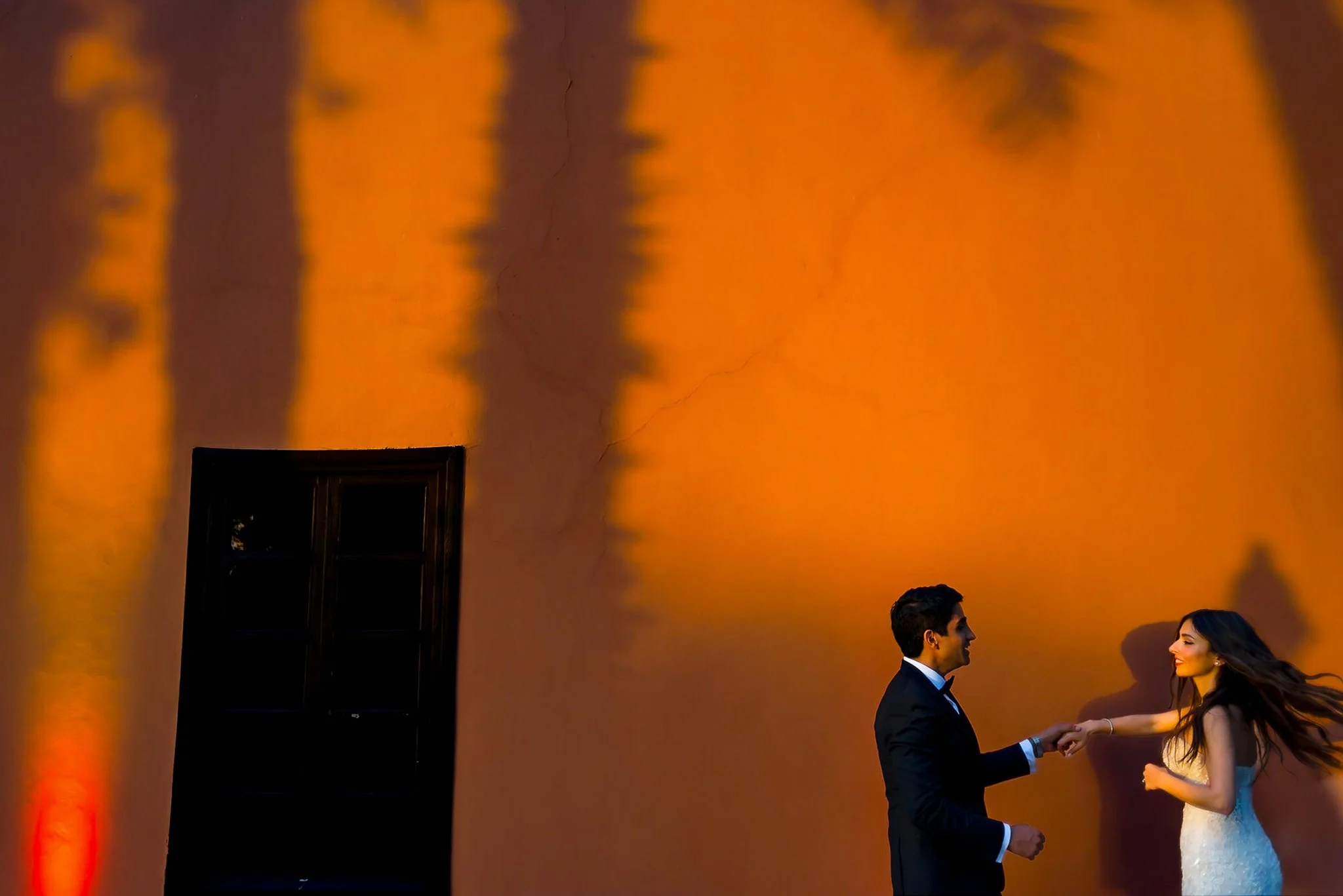 A man and a woman holding hands and dancing in front of an orange wall with shadows of trees and a window.