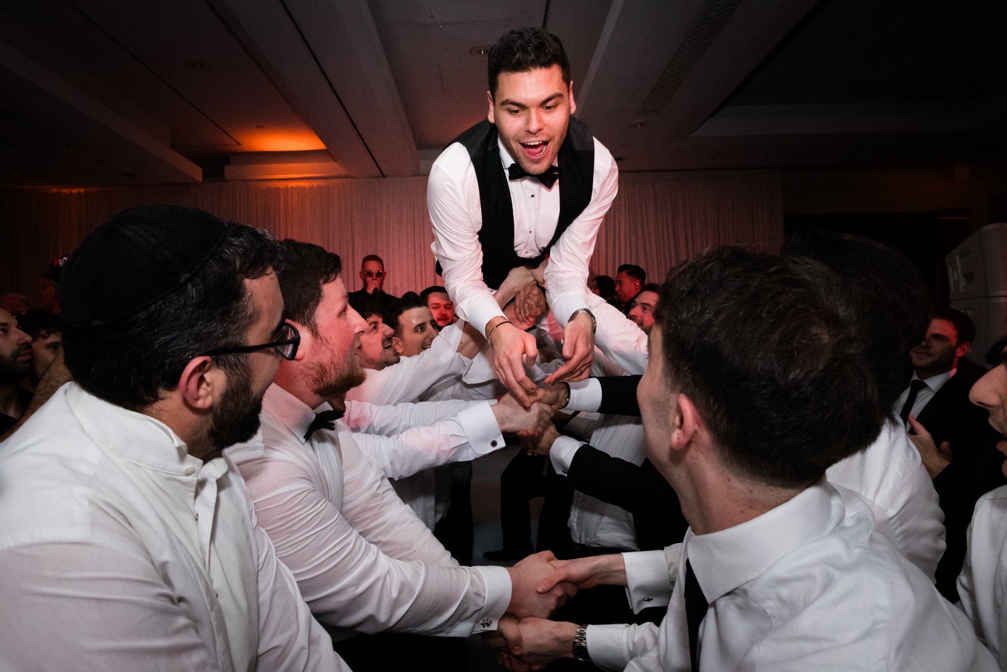 Young man in a tuxedo being lifted by a group of men in white shirts at a celebration or party.