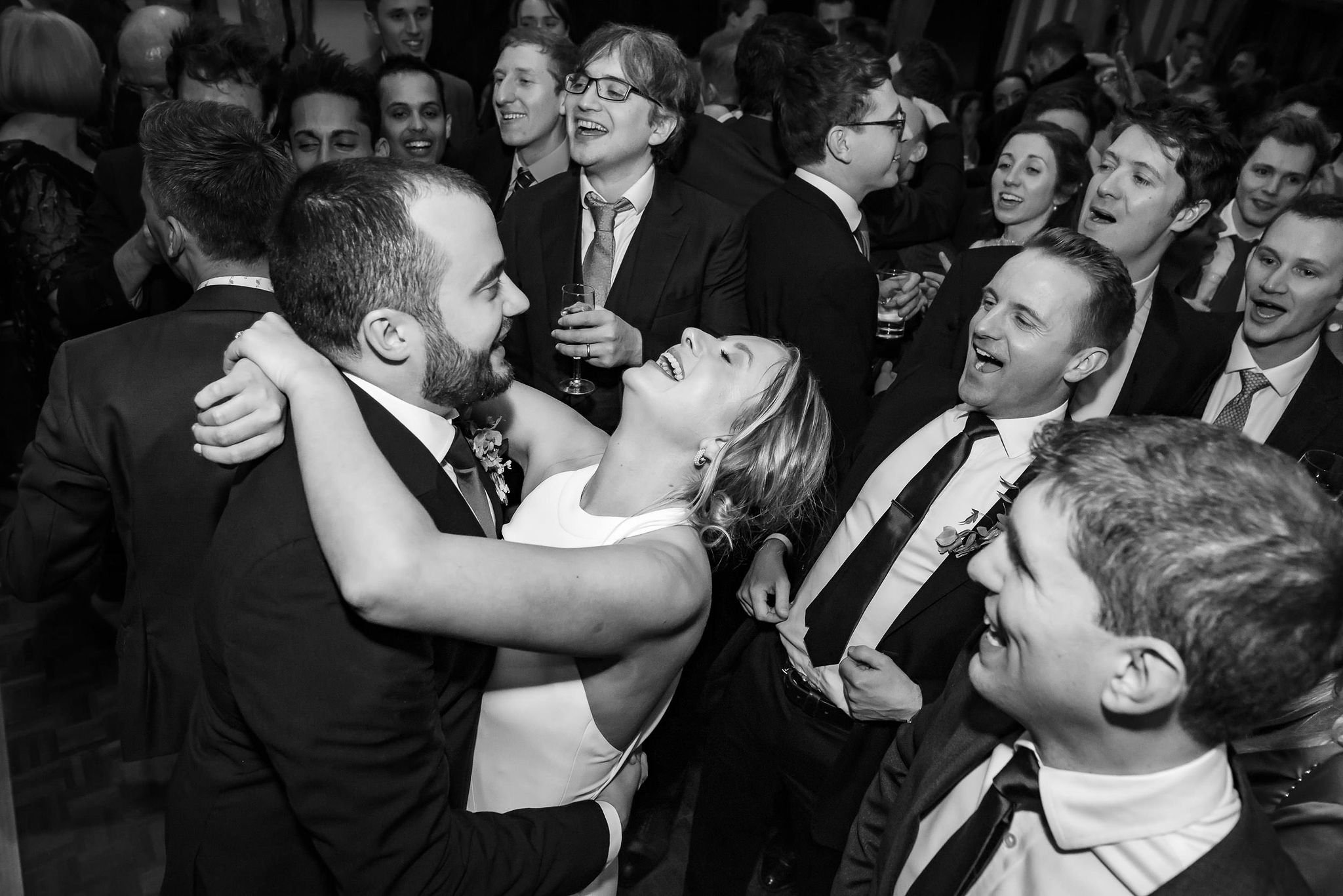 People at a celebration or wedding reception, with a bride and groom dancing and smiling in the center surrounded by guests.