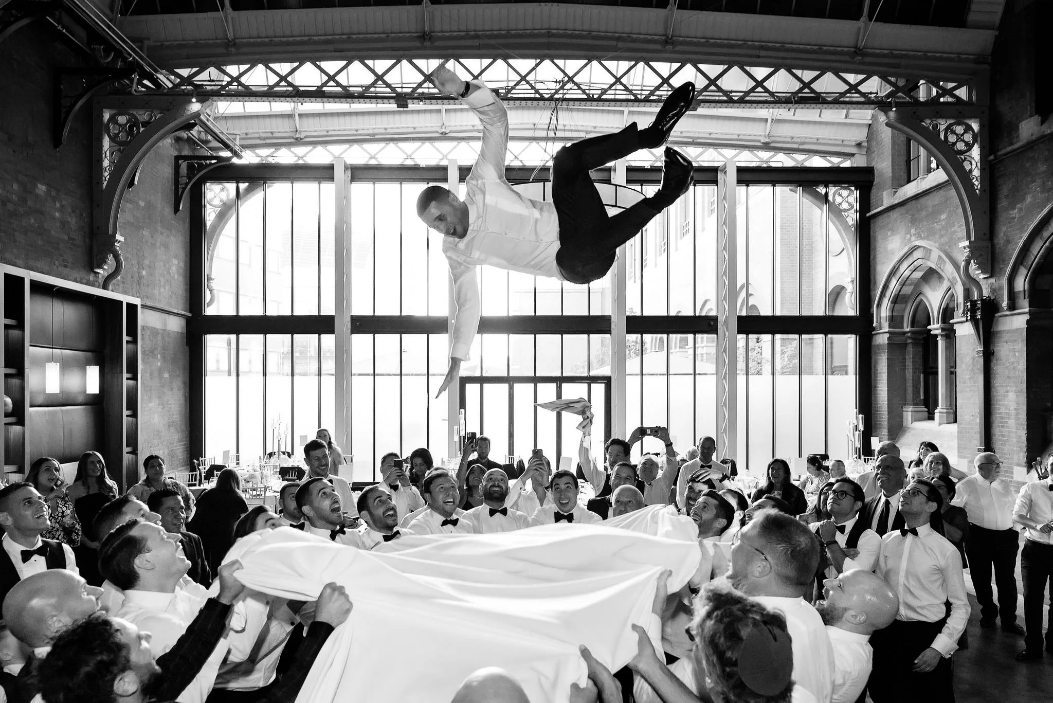 A group of people at a wedding reception lifting a groom dressed in tuxedo into the air during the reception in a decorated hall with large windows.