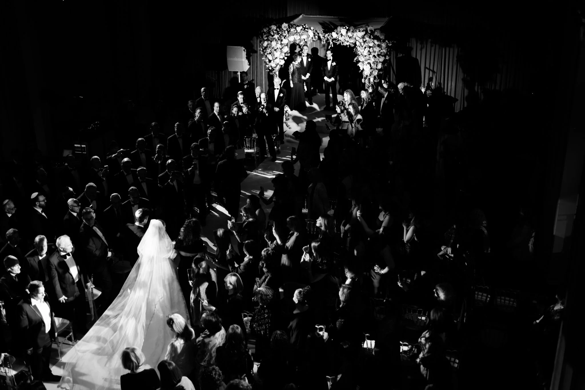 Black and white photo of a wedding ceremony with guests seated and standing, a bride with a long veil walking down the aisle, altar with floral decoration, and a couple standing at the altar with an officiant.