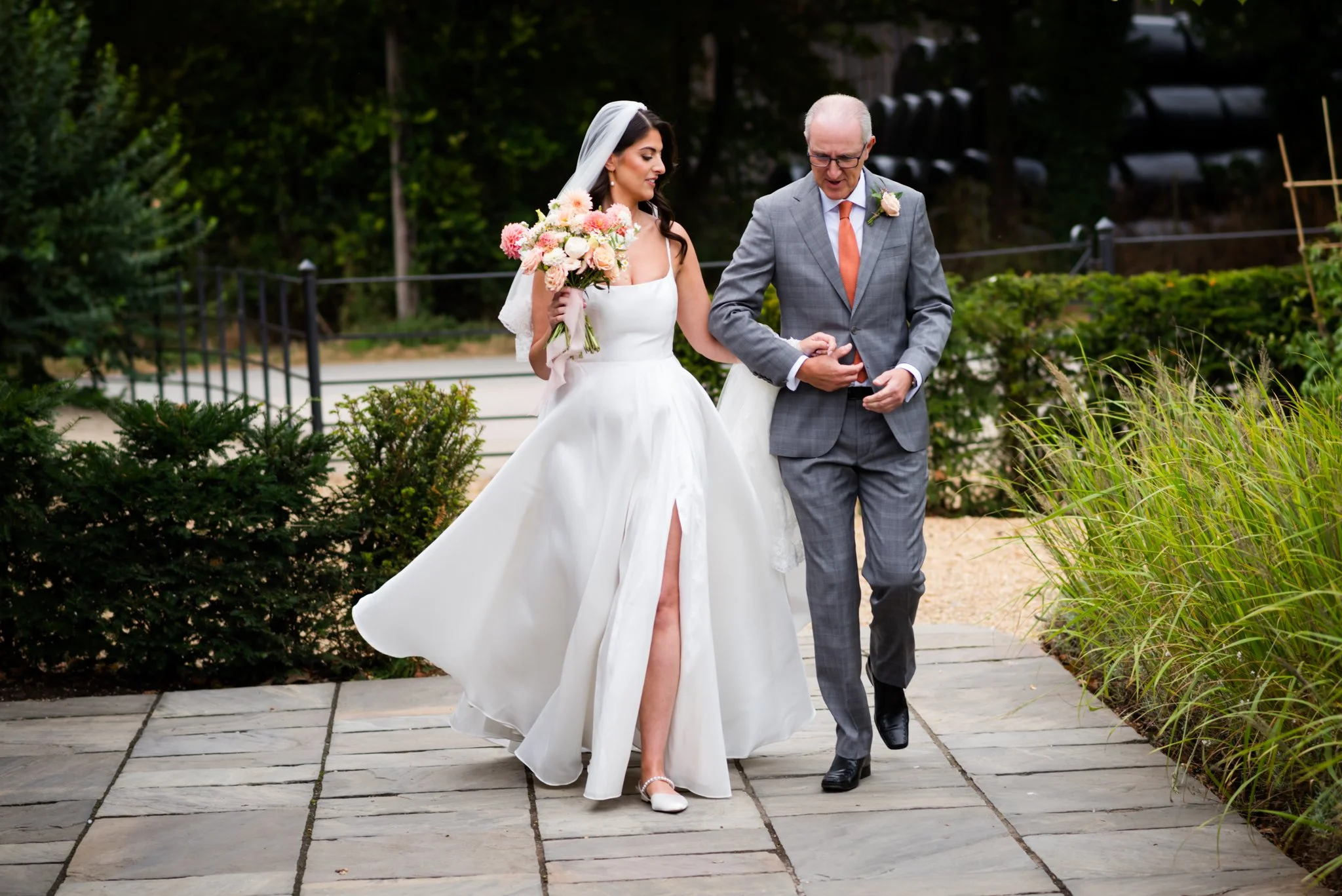 A bride wearing a white wedding dress with a slit and a veil, holding a bouquet of pink and white flowers, is walking with an older man in a gray suit, who is helping her adjust her dress, outdoors on a stone pathway surrounded by green bushes and tr