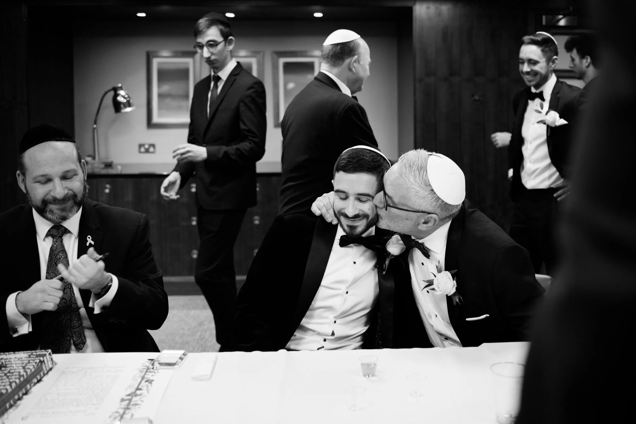 Black and white photo of a wedding celebration with men in tuxedos and yarmulkes. One man is kissing another on the cheek, both smiling. Others are standing and sitting around, some with drinks, in a warmly lit room.