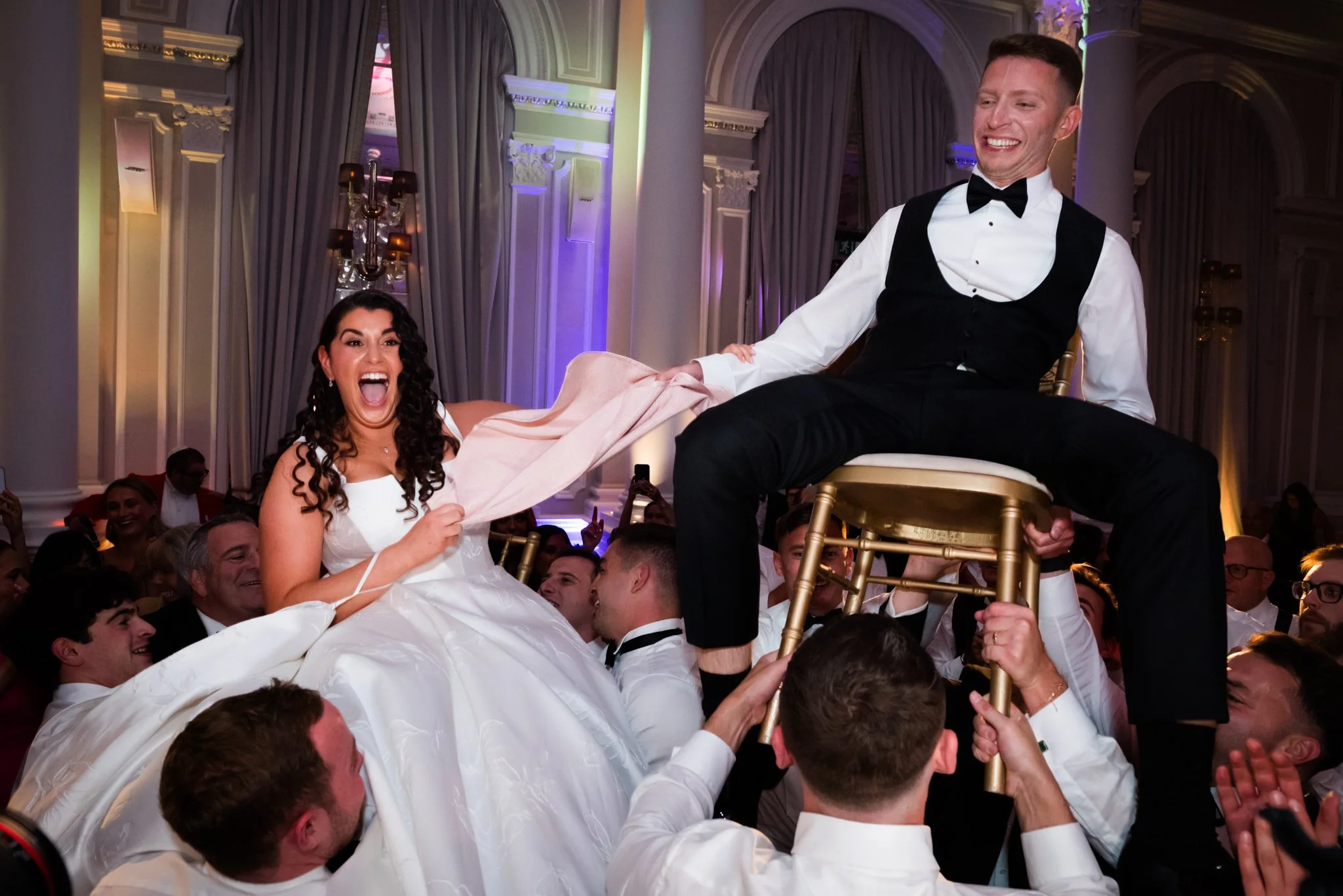 A bride and groom at a wedding celebration, with the bride sitting on the shoulders of guests and the groom sitting on a chair held by other guests. The bride is wearing a white wedding gown and appears to be laughing excitedly, while the groom is dr