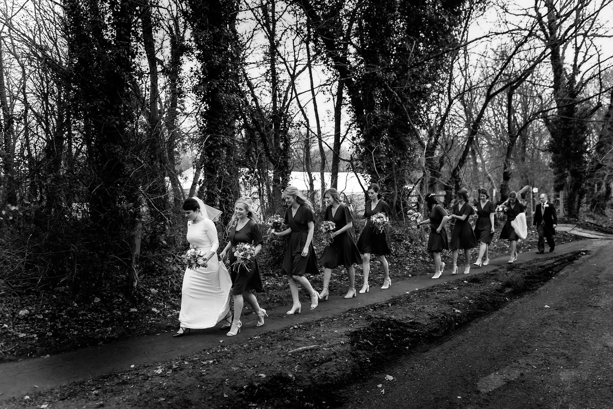 Black and white photo of a bride in a wedding dress leading a line of women in black dresses, some holding bouquets, walking along a dirt path through a wooded area, with a man in a suit following behind.