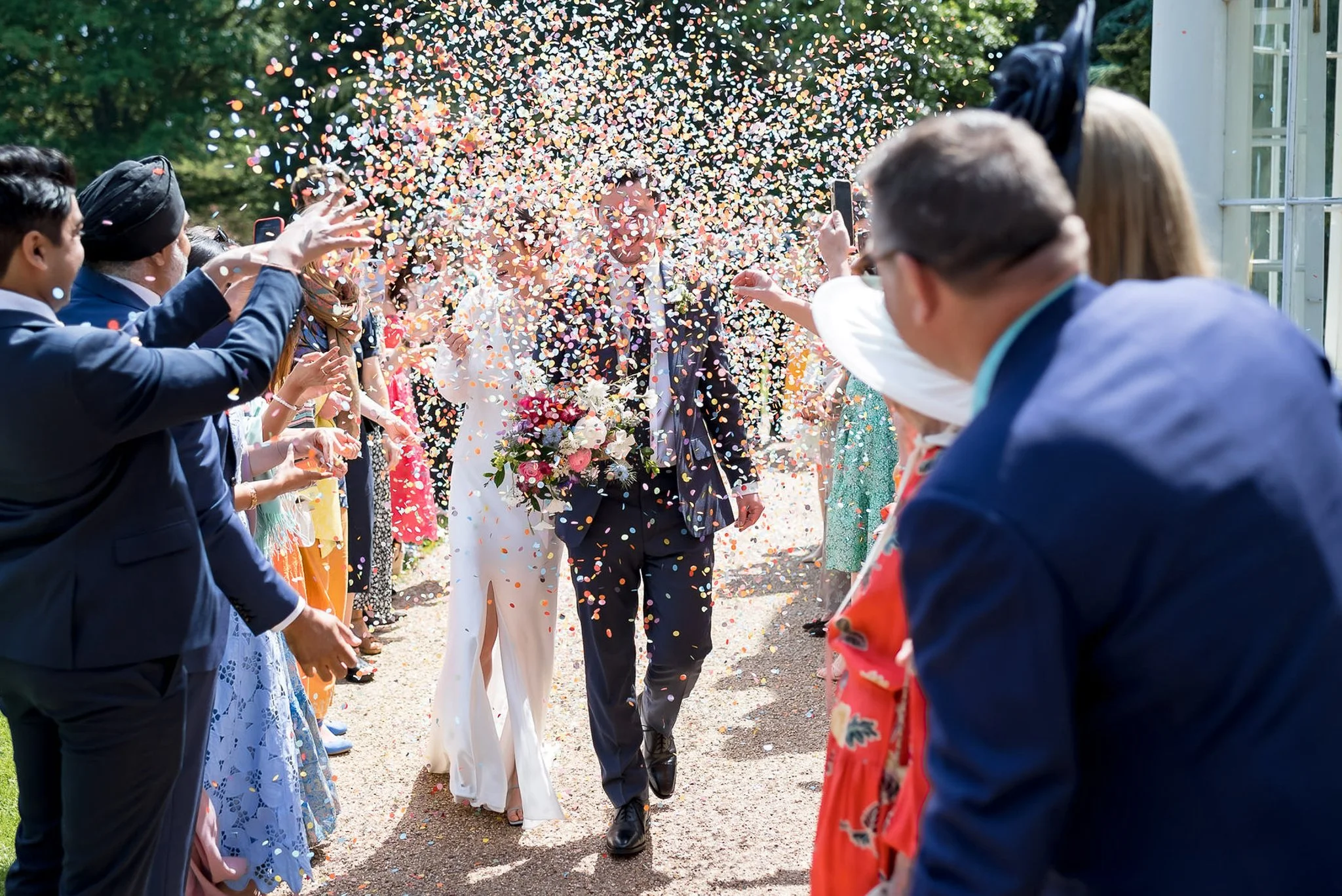 Wedding couple walking through confetti shower, surrounded by guests throwing confetti, outdoors on sunny day.