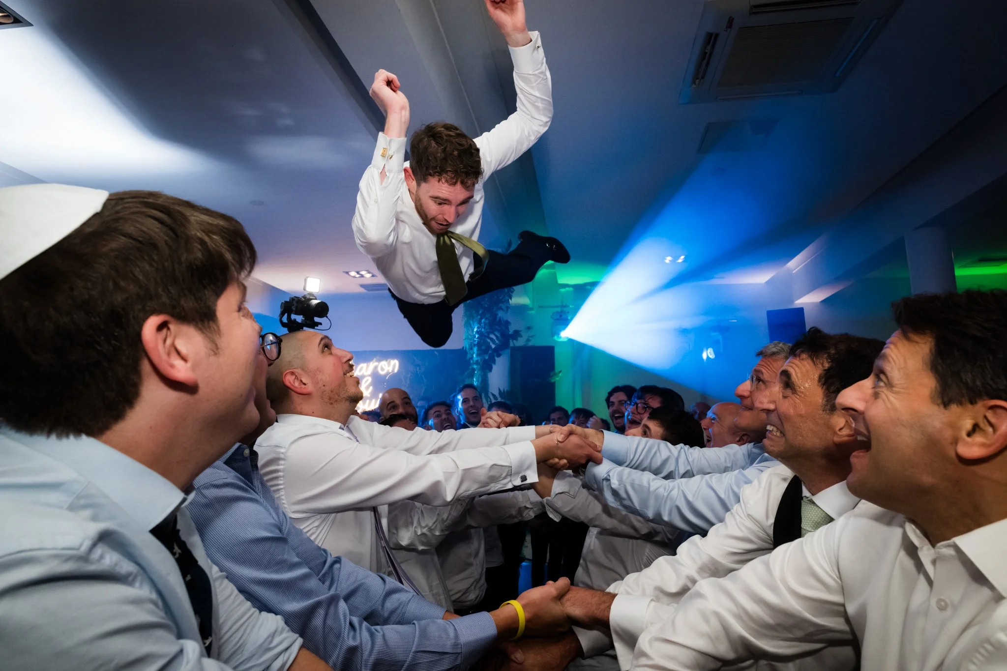 A man in a white shirt and green tie is airborne, jumping into a crowd of people at a celebration or party. The crowd is holding hands or supporting him, and everyone appears excited and happy. The setting has colorful lighting, and there is a neon s