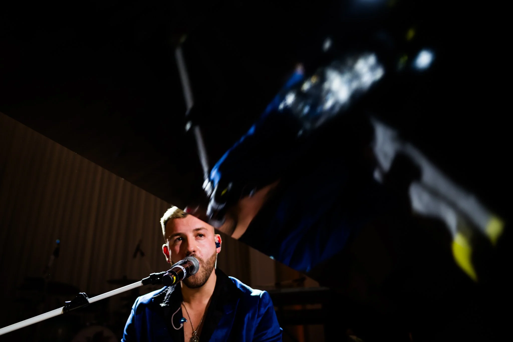 Musician singing into microphone during a live performance, with reflected image of his face and microphone on a shiny black grand piano.