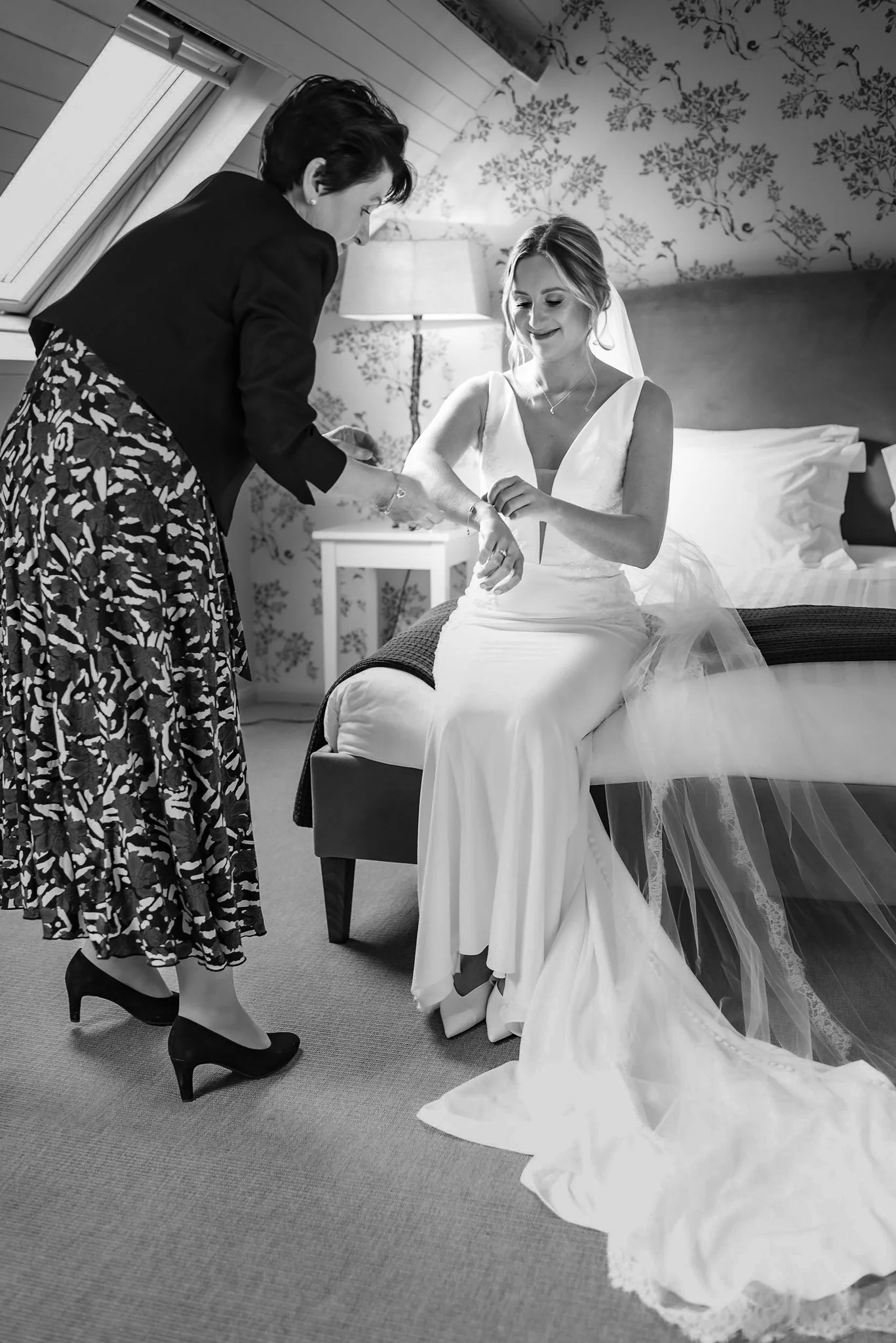 A bride in a wedding dress sitting on a bed, smiling while a woman helps her with her wristband, in a decorated room.