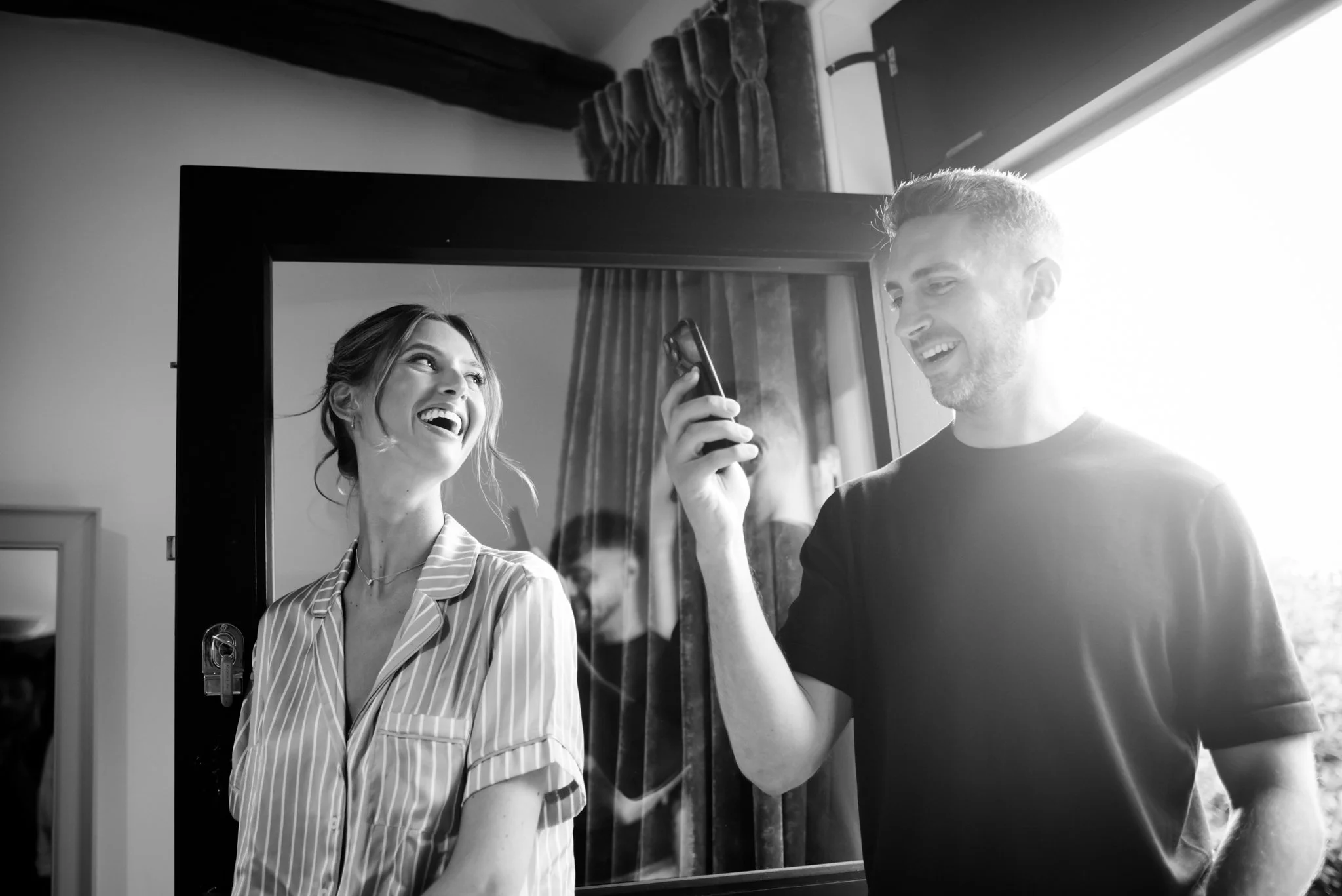 A man and woman smiling and laughing, standing in front of a mirror, with sunlight streaming in from the right.