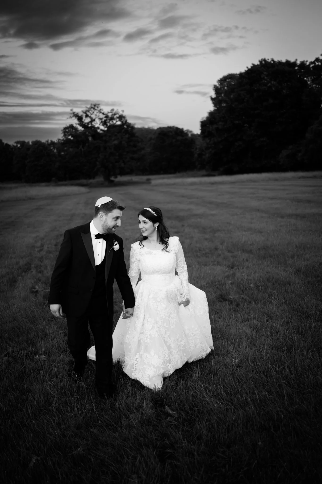 Black and white photo of a newlywed couple walking hand in hand through a grassy field, with trees in the background and a cloudy sky overhead.