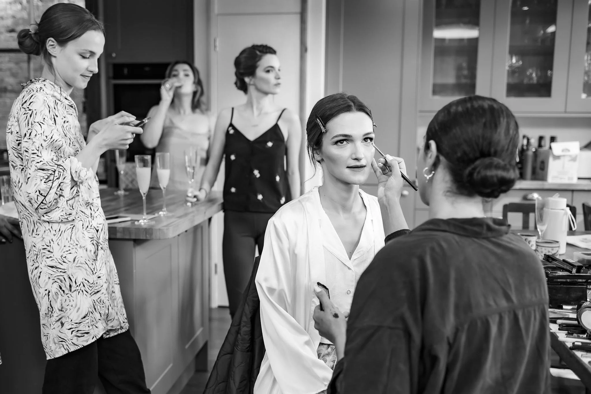 Women preparing for a photoshoot or event, with one woman having makeup applied by a makeup artist, a woman getting her makeup done, and two women in the background drinking champagne.