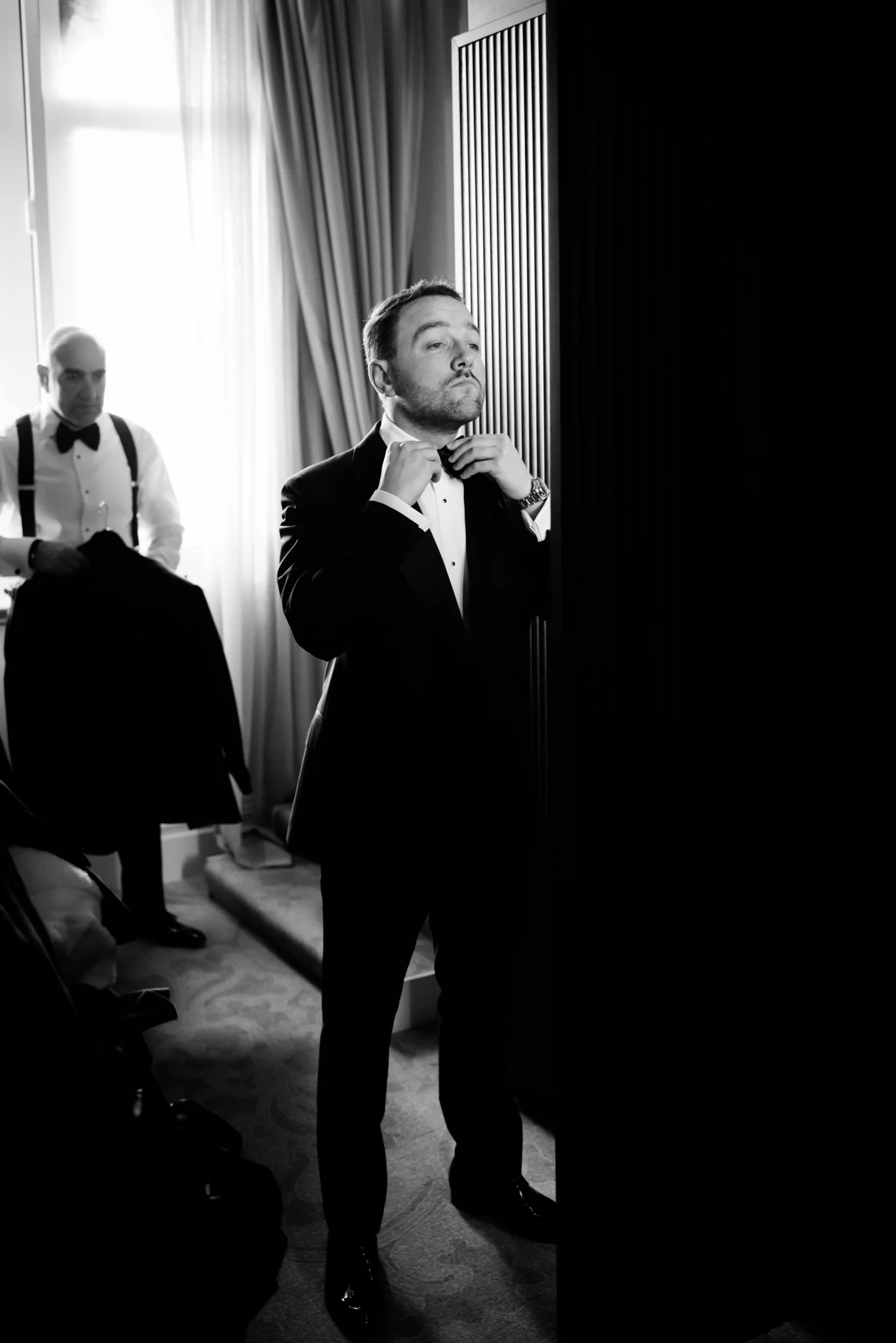 A man adjusting his bow tie in front of a mirror, dressed in a tuxedo, with another man in the background holding his jacket.