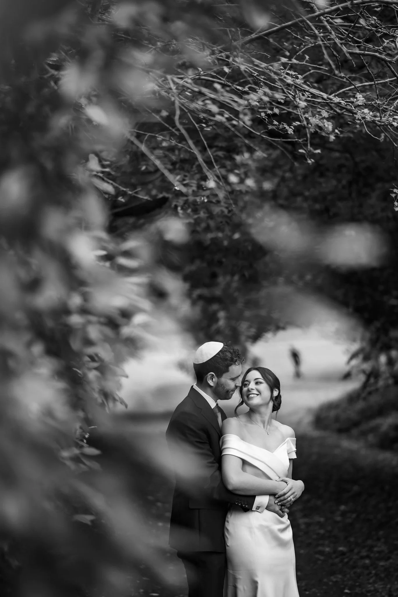 A black and white photo of a happy couple, with the man wearing a kippah, embracing and smiling at each other outdoors with trees and blurred background.