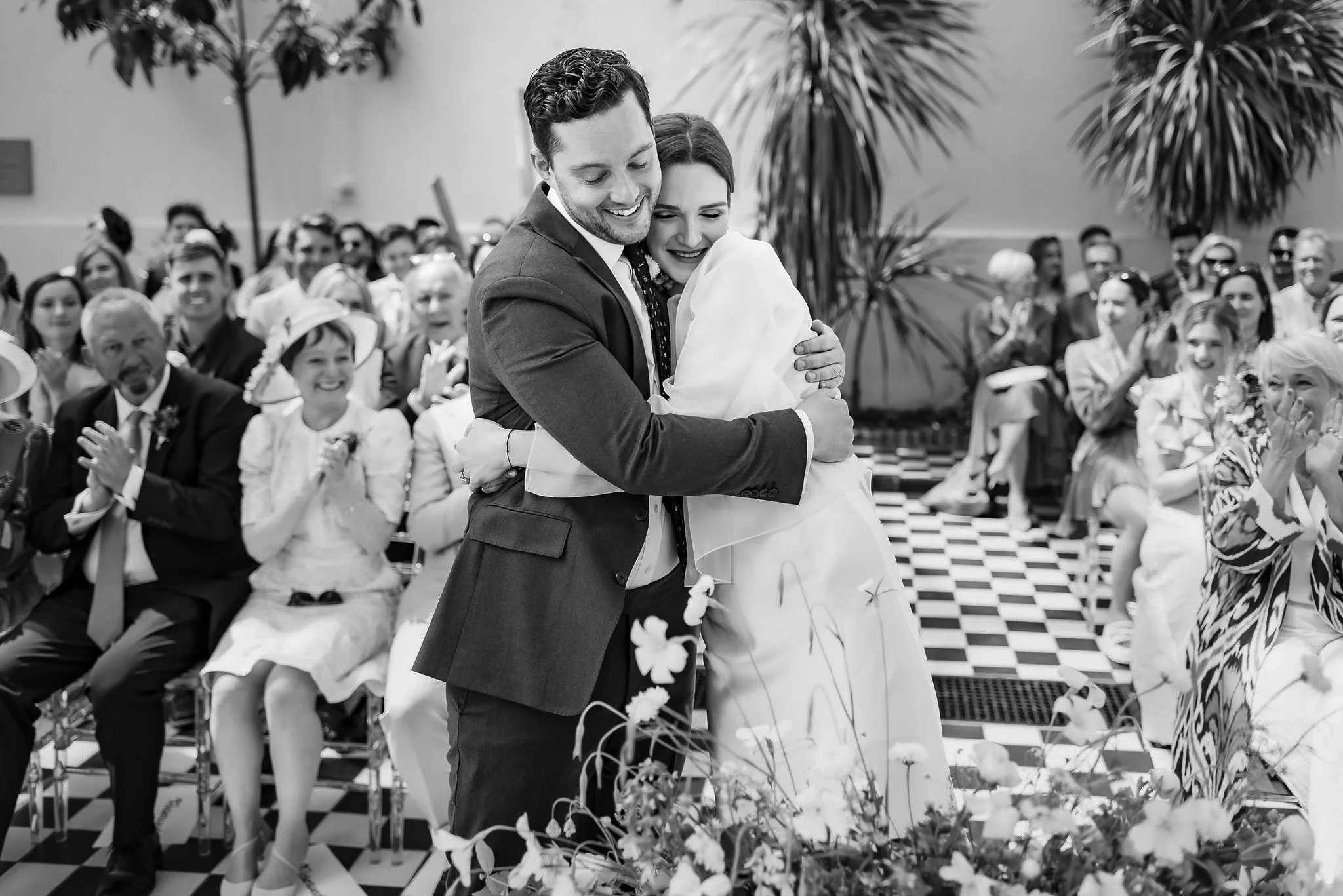 A black-and-white photo of a wedding ceremony where a groom and bride are hugging. The groom is wearing a dark suit and the bride is in a wedding dress. Guests are seated in the background, smiling and clapping, with some wearing hats and sunglasses.