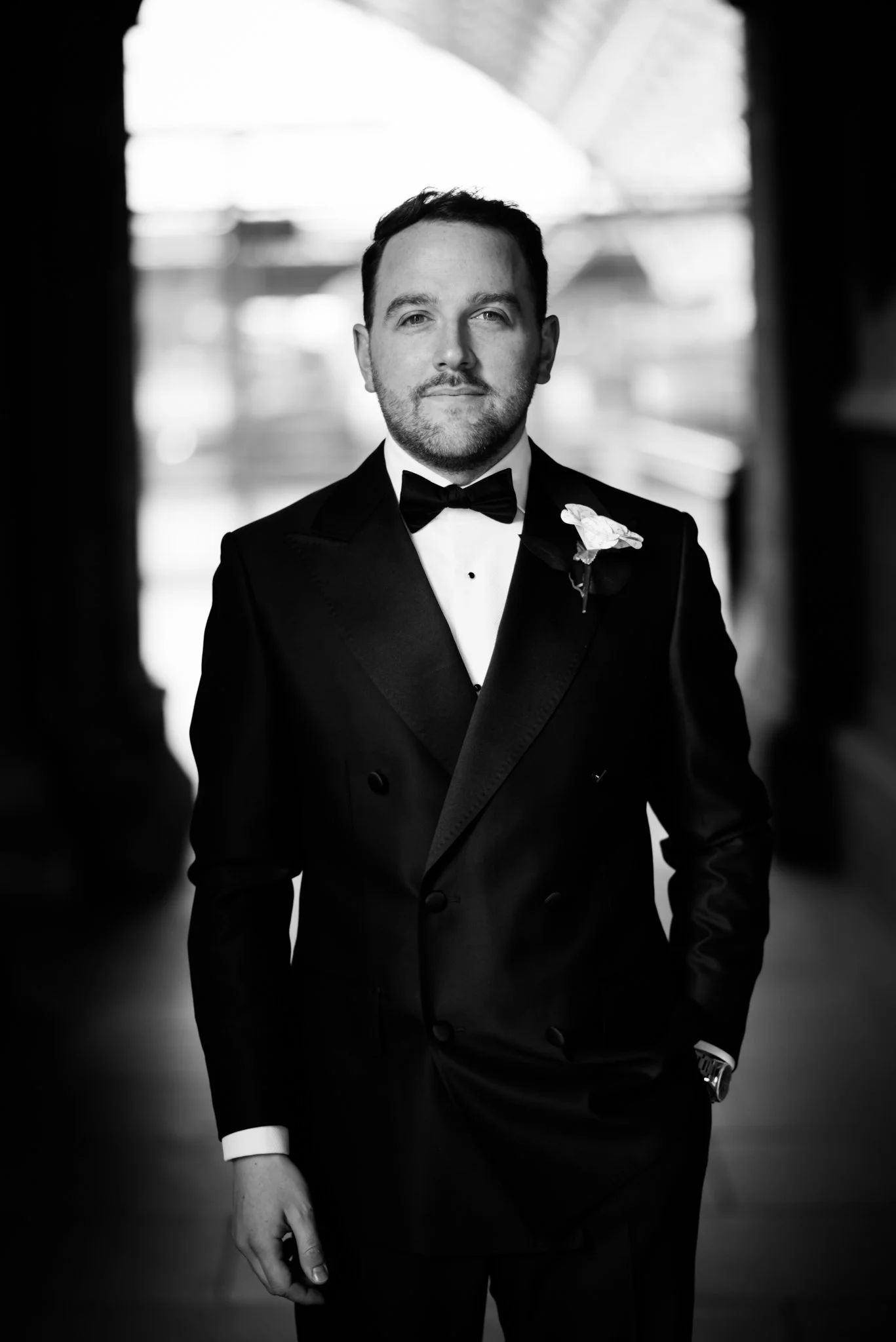 A man dressed in a tuxedo with a bow tie, standing indoors with blurred background, looking confidently at the camera.