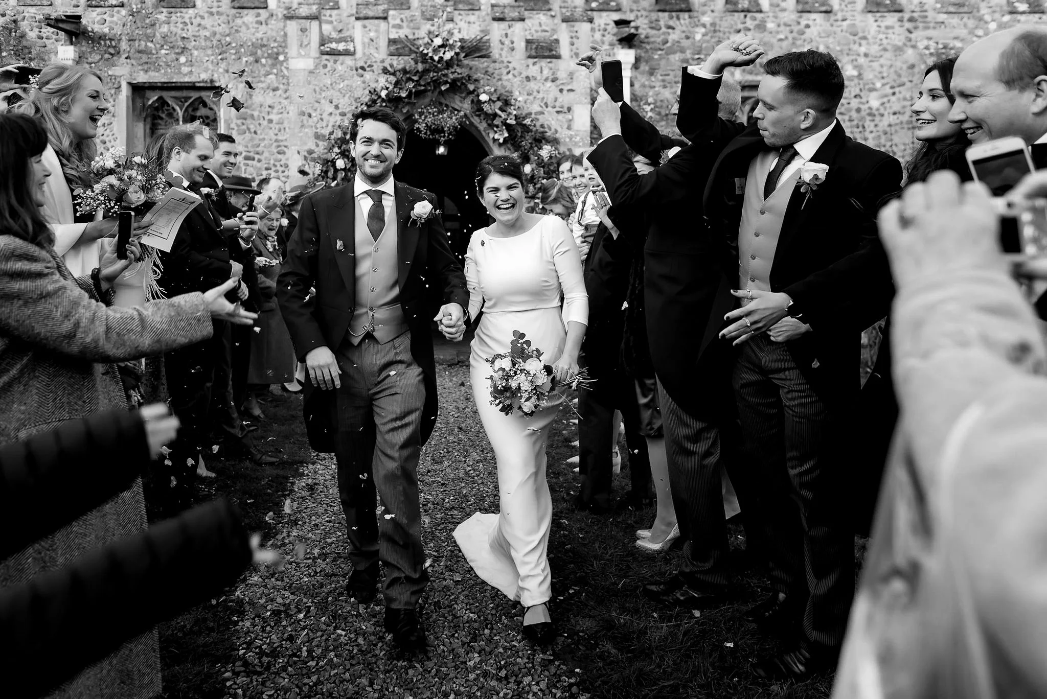 A black and white photograph of a wedding celebration outside a stone church. The bride and groom are walking hand in hand, smiling, surrounded by friends and family throwing celebratory confetti. The bride is holding a bouquet, and guests are captur
