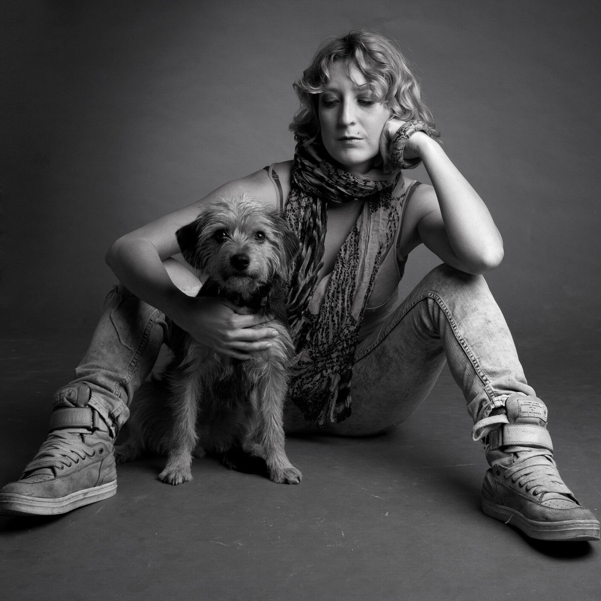 Black and white photo of a woman sitting on the ground with her knees bent, holding a small scruffy dog close to her chest. The woman has curly hair, wears a scarf, a sleeveless top, and sneakers. The dog looks directly at the camera.