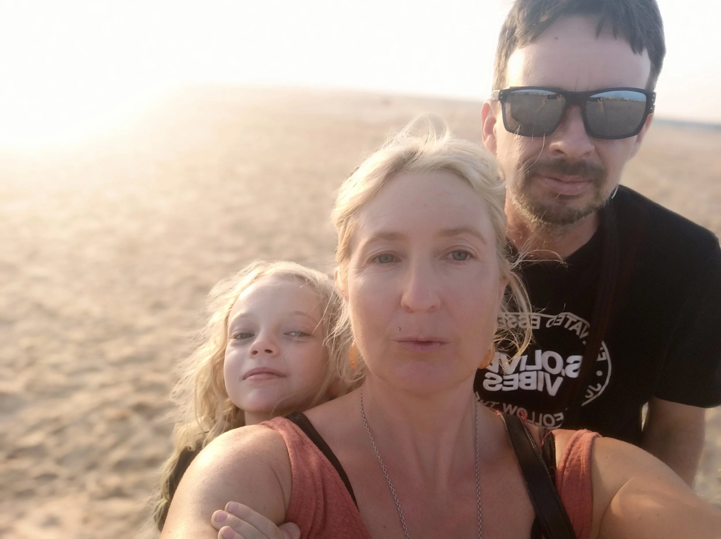 A family of three taking a selfie on a beach during sunset, with the ocean in the background. The woman is in the foreground, wearing a reddish top, with a young girl hugging her from behind. The man is in the background, wearing sunglasses and a black shirt.