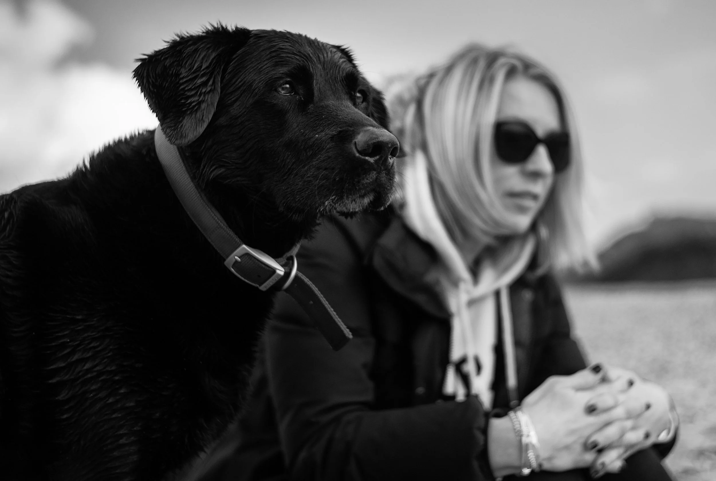 A woman with blonde hair, wearing dark sunglasses, a hoodie, and a jacket, sitting outdoors with her hands clasped.