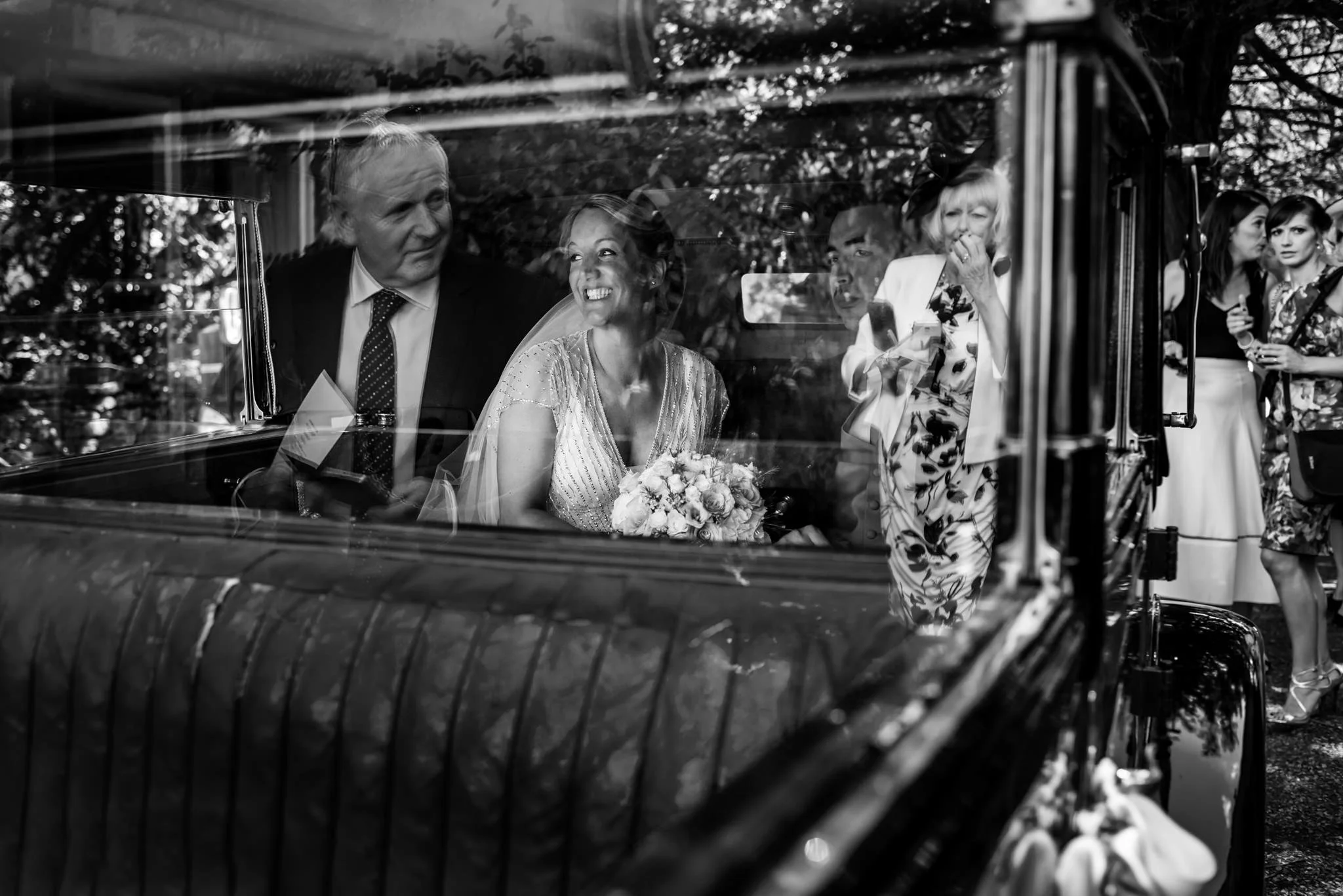 A black and white photo taken through a car window, featuring a smiling bride holding a bouquet inside a vintage car, with several women standing outside the car, some talking and others observing, in a garden setting.