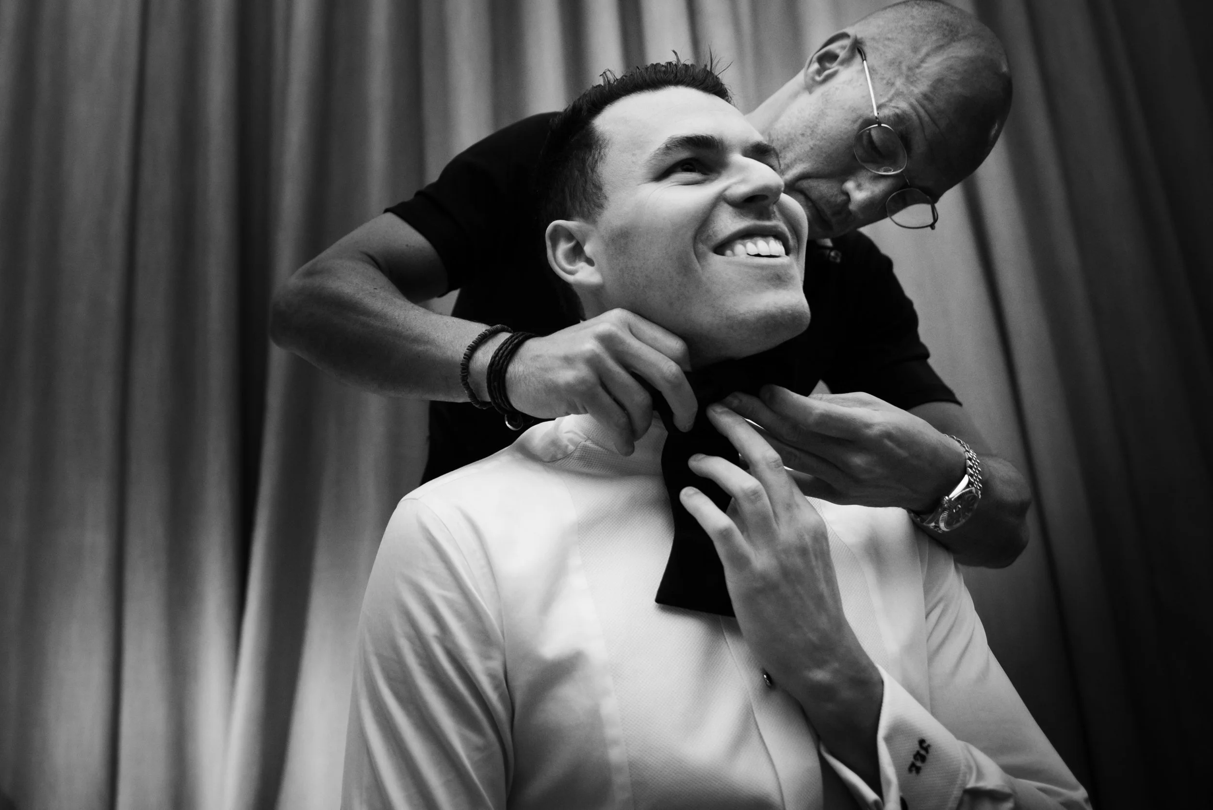 A young man in a white dress shirt is being assisted by an older man, who is helping him adjust a black bow tie and is leaning close to him. The younger man is smiling happily while the older man, wearing glasses, is focused on helping him. They are 