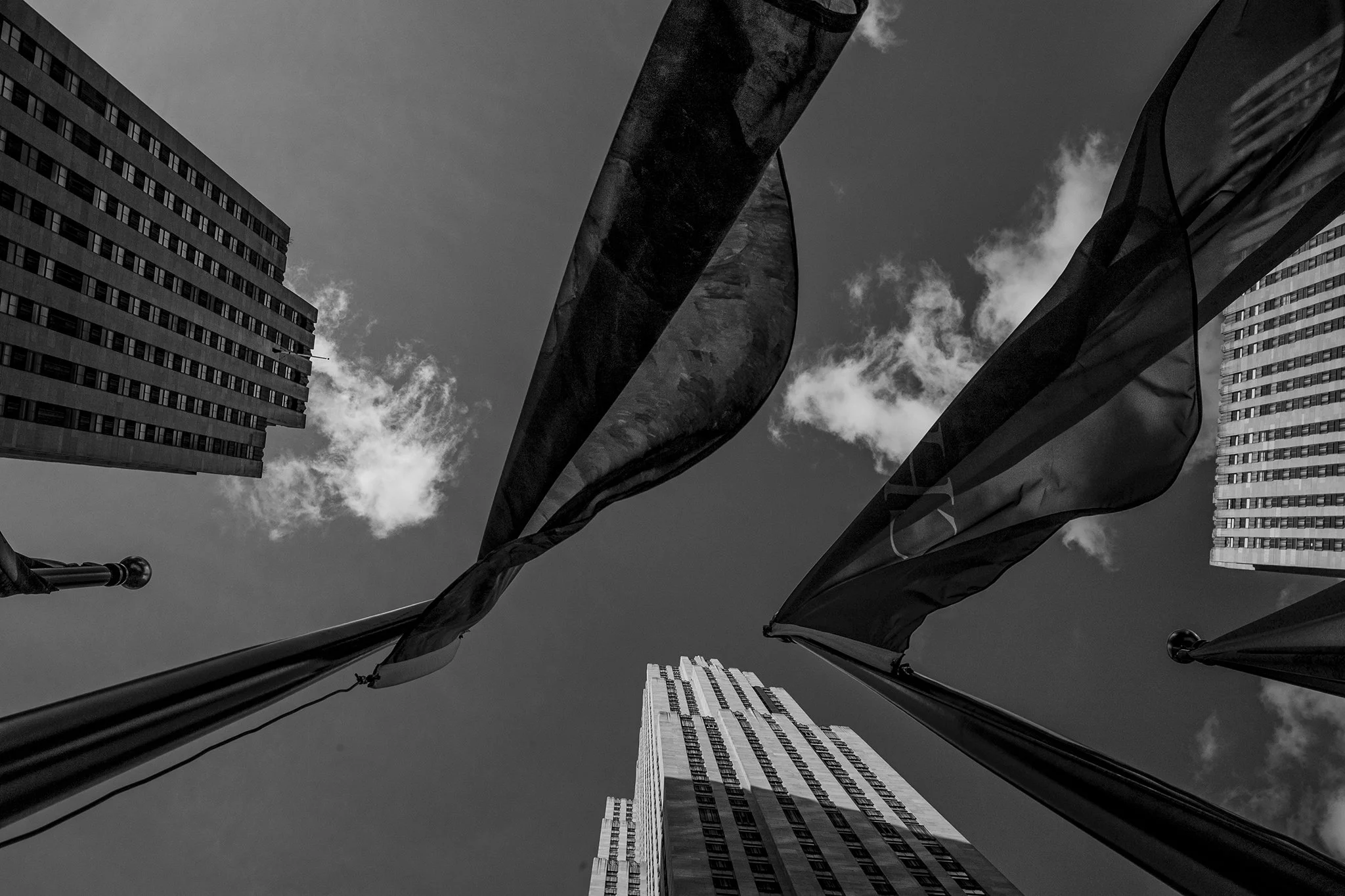 View of flagpoles with flags in an urban setting, seen from below with tall buildings and a partly cloudy sky in the background.